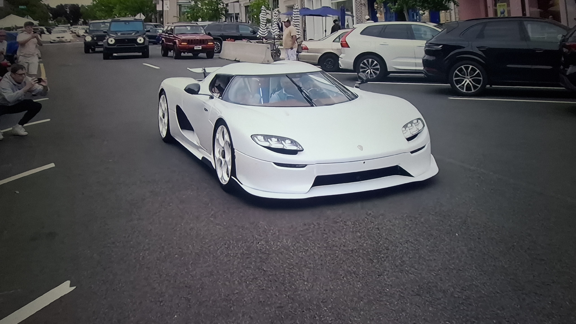 A white Koenigsegg supercar, likely a CCX or CCXR, drives slowly down an asphalt street, serving as the central focus of the image. Its sleek, aerodynamic design and white finish stand out, with large white multi-spoke wheels visible. Two occupants can be faintly seen inside the cabin.
In the mid-ground, a black Mercedes-Benz G-Wagen and a red Jeep Cherokee XJ are visible, traveling in the same direction or parked. Further in the background, a white Volvo XC60 and a black Porsche Cayenne (or Macan) are parked alongside other various vehicles, indicating a street or urban setting.
Several people are present, observing the cars. In the left foreground, a man in a grey hoodie and white sneakers crouches, actively taking a photograph or video of the passing supercar with his phone. Other spectators are visible in the background near parked cars and buildings, some under blue and white umbrellas, suggesting a casual gathering or event.
The scene takes place during the daytime, under bright, possibly overcast, conditions. The road surface is dark asphalt with white painted lines. No discernible text is visible on any of the vehicles or in the environment. The overall atmosphere suggests a car show, rally, or enthusiast event where people are gathered to admire and document high-performance automobiles.