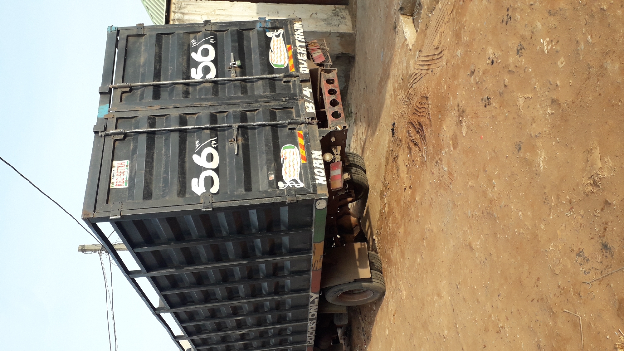 The image depicts a truck parked on a dirt lot in Nkpor, Nigeria. The truck consists of two stacked containers. The containers are dark with the number "56" painted on the side in white. The container has “RBL-967XH” printed on a rectangular plate on the side. The truck also has a sign that says "HORN" and an illustration of what appears to be white plumes. The ground is brown, dirt and some debris scattered about. A building and utility pole with electrical lines are visible in the background. The sky is visible in the upper-left corner.
