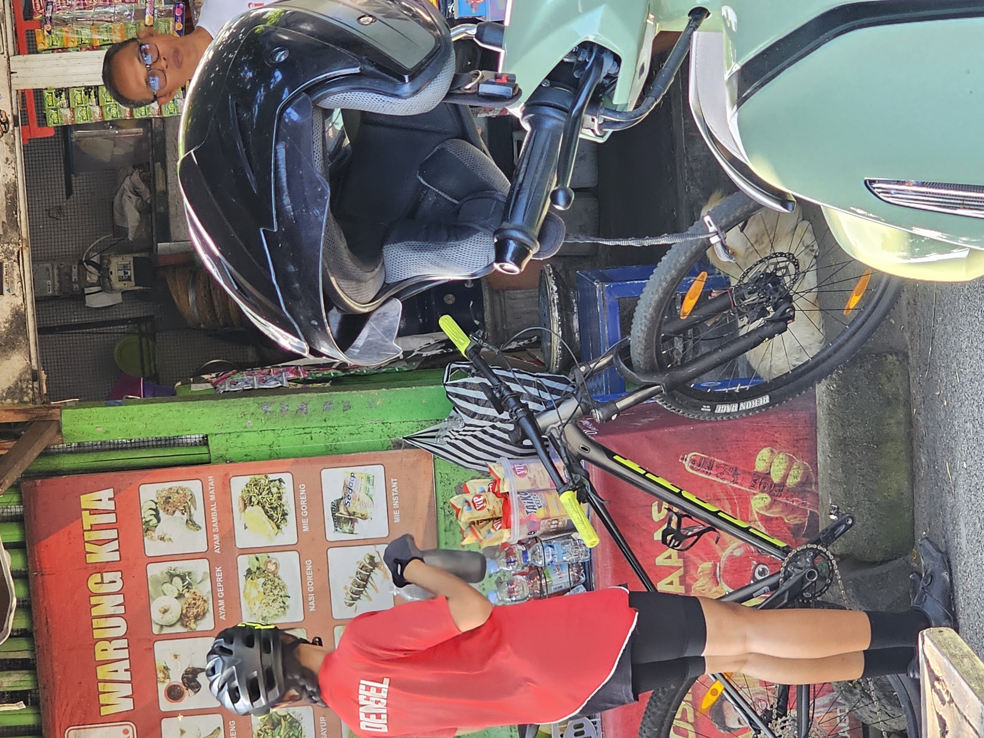 The image depicts a street scene in Denpasar, Indonesia. A person wearing cycling gear (red jersey, black shorts, gloves, helmet) stands next to a bicycle. Behind the person is a "Warung Kita" sign featuring Indonesian dishes like Ayam Geprek, Ayam Sambal Matah, Nasi Goreng, Mie Goreng, and Mie Instant. A motorcycle and a black helmet are also visible, along with a person wearing glasses. Bottles of water and snacks are on the shelf.