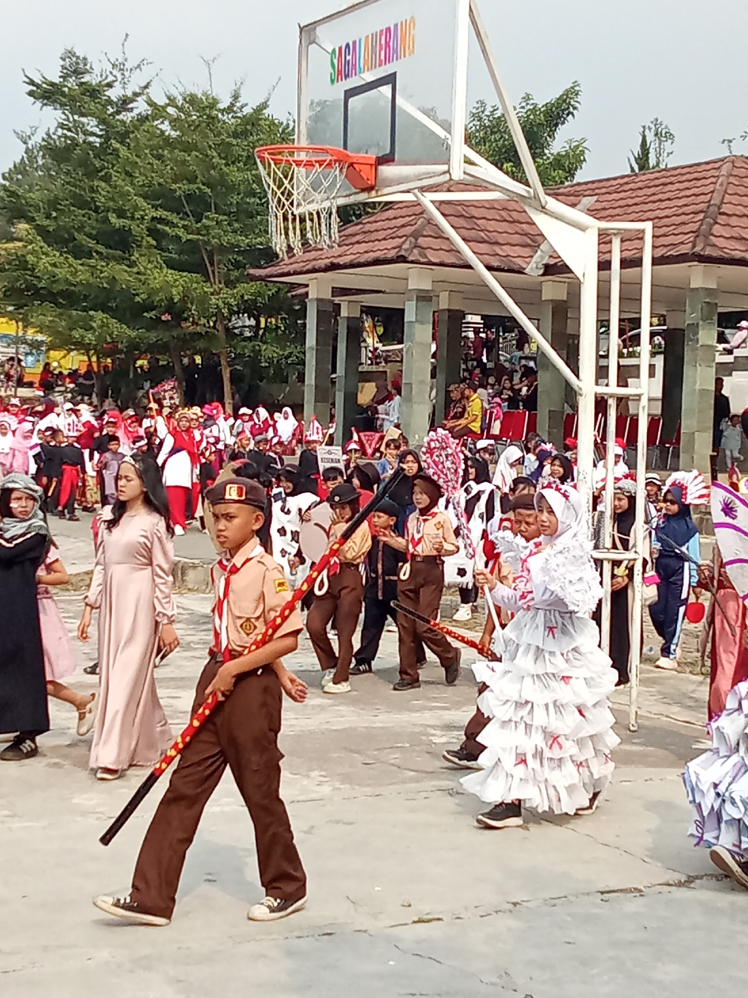 In Subang, Indonesia, a group of children participates in a parade. The scene is set outdoors with a basketball hoop and a covered pavilion in the background. Several children are dressed in scout uniforms, while others wear festive outfits, including a girl in a white, ruffled costume. Some children carry decorative staffs. The ground is paved with concrete, and trees are visible in the distance. A sign on the pavilion reads "SAGALHERANG".
