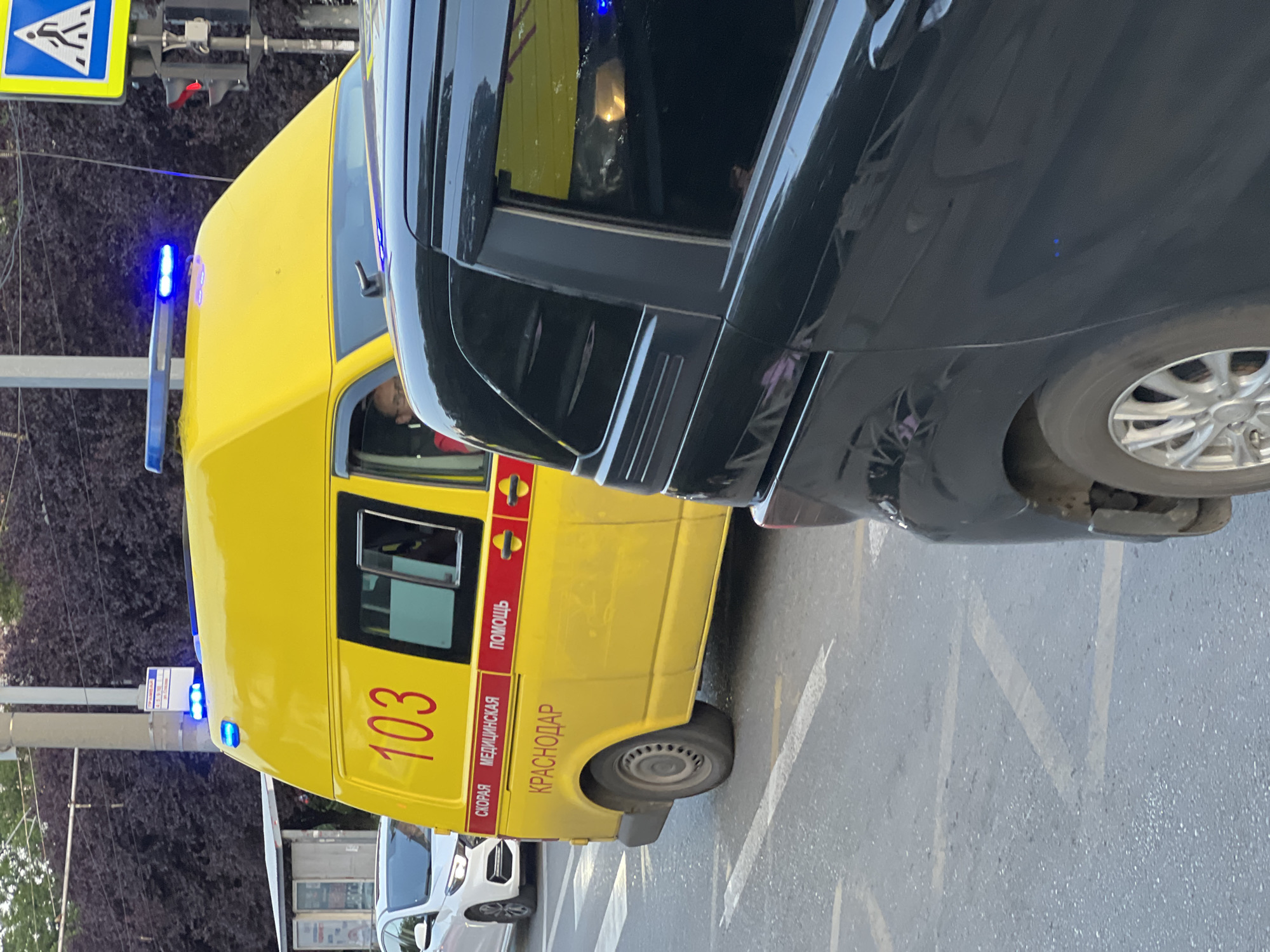 The image shows a yellow ambulance with blue flashing lights on its roof, parked on a city street in Krasnodar, Russia. The ambulance has the number "103" and text in Russian indicating "Ambulance Medical Aid" and "Krasnodar" written on its side. A person is visible through the driver's side window, wearing glasses and a red garment. A black car is partially obscuring the view of the ambulance from the right side of the frame. In the background, a white car is seen driving away. The scene appears to be an urban environment with trees and a traffic light visible.