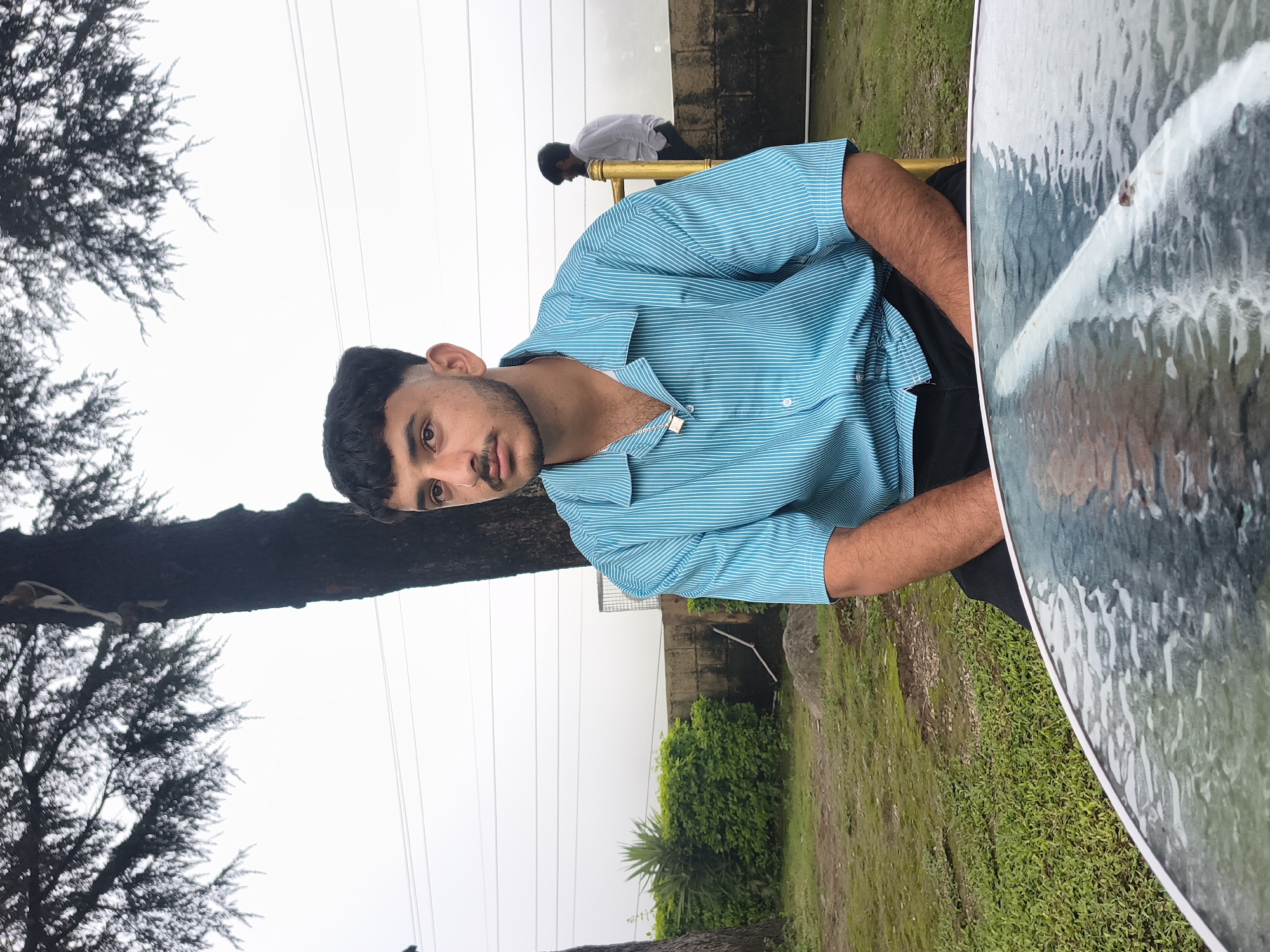 A young man with dark, short hair and a mustache sits on a golden chair, facing slightly away from the camera. He is wearing a turquoise and white striped short-sleeved collared shirt and black pants, with a silver chain around his neck. Behind him, a second man in a white shirt stands near a low stone wall. The scene appears to be an outdoor seating area, possibly a cafe or restaurant, with a glass-topped table in the foreground. Lush green grass and trees, including a large tree with intricate branches, surround the area. The background is dominated by a pale, overcast sky and power lines. The location is identified as Khaniara, India.