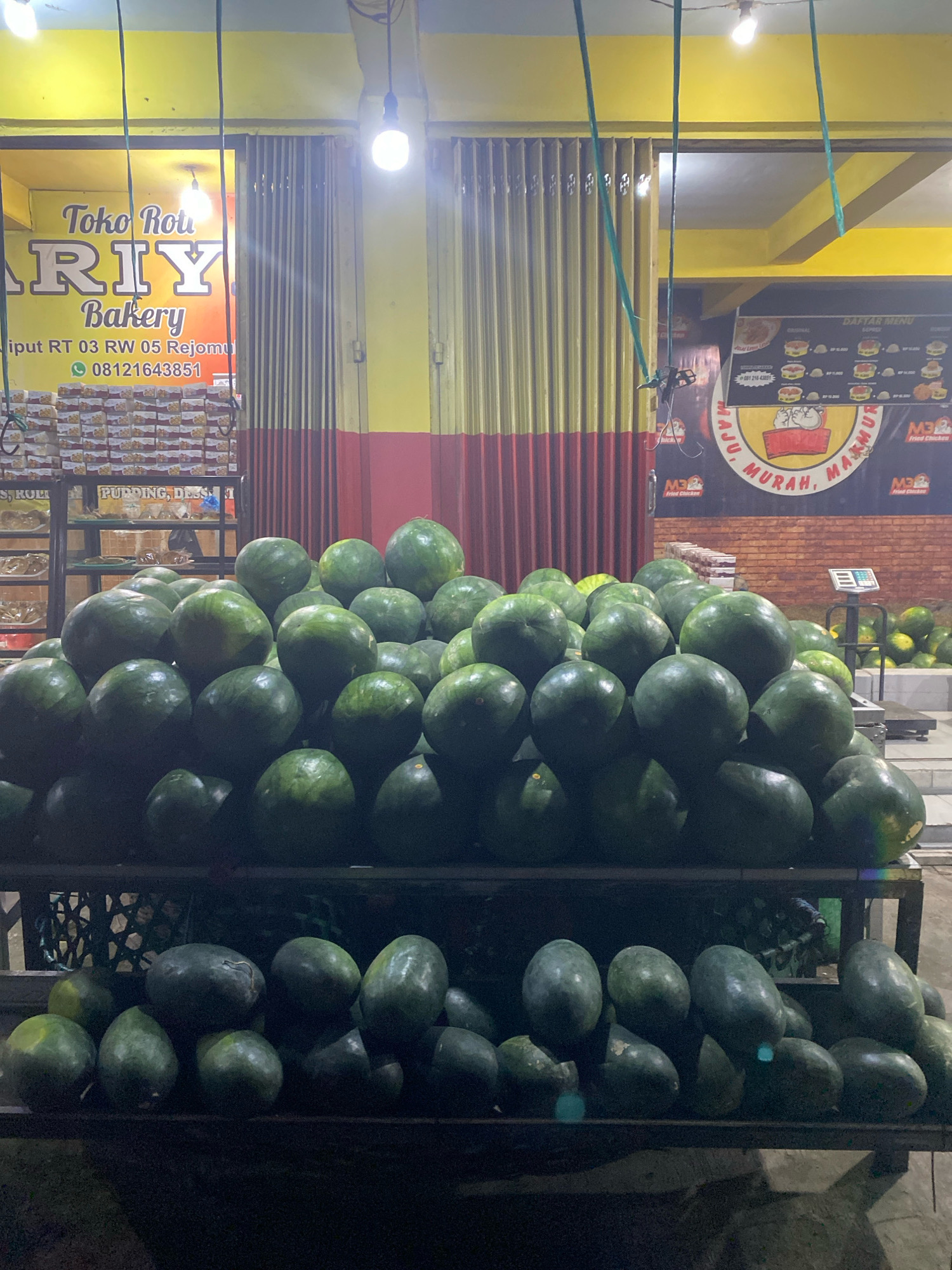 The image depicts an outdoor market stall selling watermelons in the city of Kediri, Indonesia. The stall features several racks overflowing with a large quantity of watermelons, predominantly dark green in color. Some watermelons are stacked in neat pyramids, while others are more loosely arranged. In the background, there are signs for a bakery, "Toko Roti RIY Bakery," with a phone number visible. Further back, a menu board for "M3 FRIED CHICKEN" is displayed. Shelves stocked with packaged goods, possibly baked items, are also present. The lighting suggests it is either evening or a covered area with artificial lights. The ground appears to be concrete. There are no people prominently visible in the scene, but the presence of a weighing scale suggests a commercial transaction is likely occurring or has recently occurred.