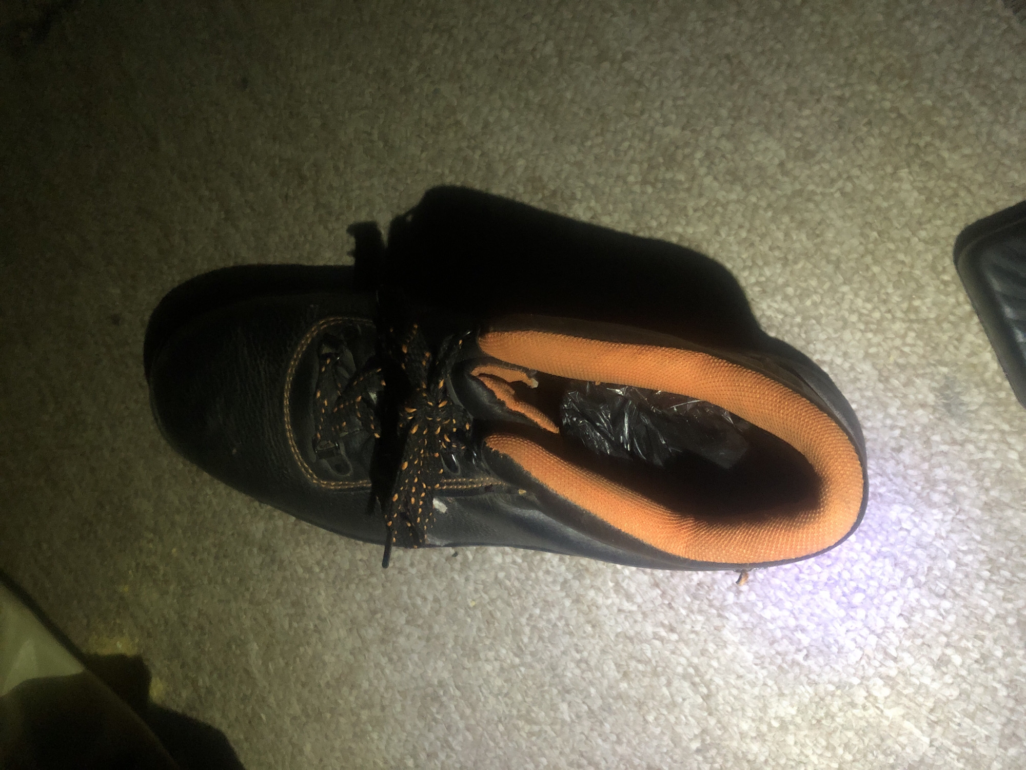 An overhead shot shows a black and orange work boot on a textured carpeted floor. The boot is laced with black and orange speckled laces. The top cuff of the boot is lined with orange padding. Inside the boot, a crumpled black plastic bag is visible. A light source, possibly a flashlight, casts a purple-blue glow on the carpet near the toe of the boot. The lighting creates strong shadows, suggesting a dimly lit environment. The background is out of focus and appears to be a floor or ground surface. There is no indication of the city of Jalingo, Nigeria, within the image.