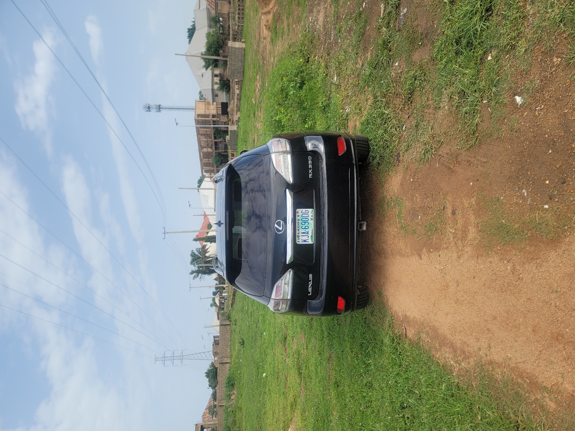 A black Lexus RX 350 is parked on a dirt road in an unpaved lot. The car is facing away from the camera, and its license plate reads "KJA-690DG". The car is surrounded by tall grass and some scattered debris. In the background, there are several buildings, a power line tower, and a cell phone tower. The sky is partly cloudy. The location is likely Jos, Nigeria, given the license plate format.
