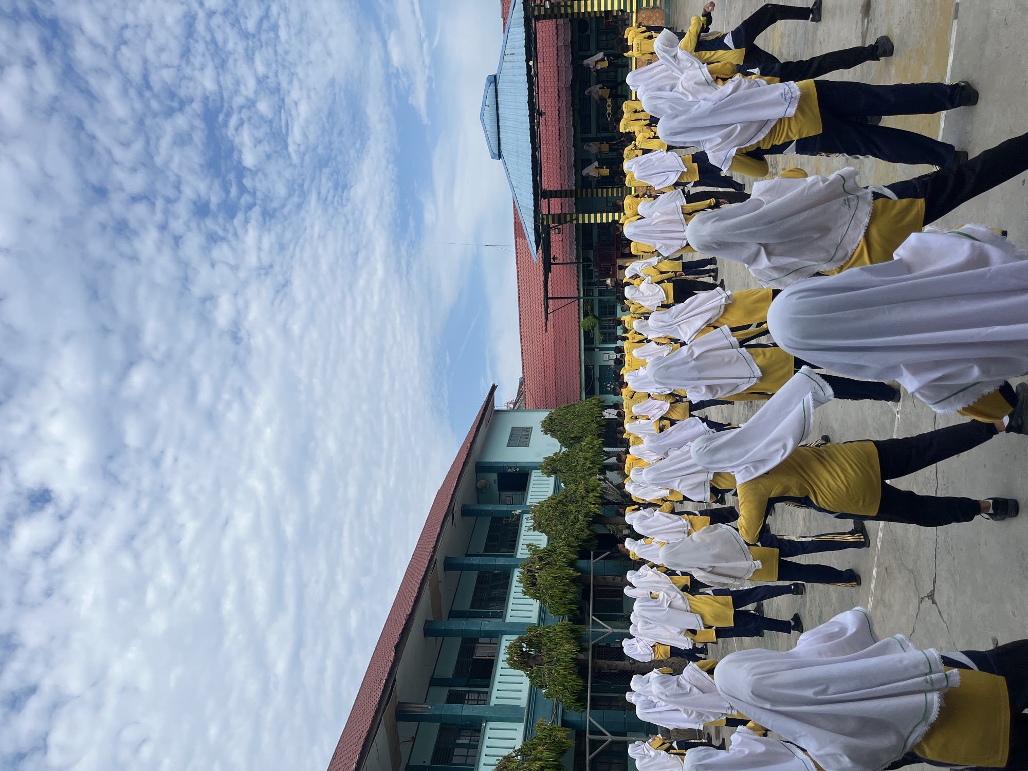 A group of students in yellow shirts and white hijabs are participating in a physical activity outdoors on a paved schoolyard. They appear to be in a coordinated movement, possibly a dance or exercise routine, facing towards a raised stage area with a blue roof. The background features a two-story school building with teal walls and a red roof, and lush green trees lining the front of the building. The sky is partly cloudy with a bright blue sky peeking through. The image was taken on the grounds of a school in Lhokseumawe, Indonesia, likely during a morning assembly or a special event.