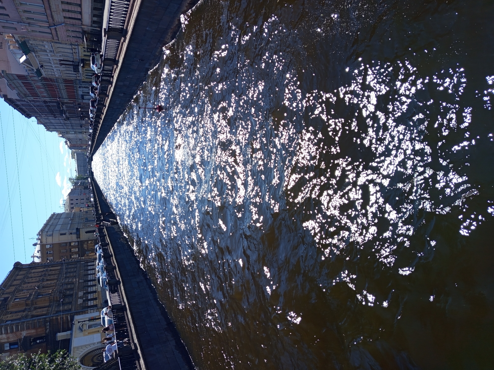 A high-angle, vertical shot captures a wide canal in Saint Petersburg, Russia, on a sunny day. The canal's dark water is illuminated by intense sun glare, creating numerous bright, shimmering highlights across its surface. On the left bank, a dark stone embankment is topped by a black metal fence. Beyond the fence, a sidewalk is lined with trees and a row of cars parked along the street. People are visible walking on the sidewalk. The buildings lining the street are tall and multi-story, with architectural styles consistent with historic Saint Petersburg. The right bank of the canal also features a stone embankment and a black metal fence. Beyond the fence, cars are parked along the street, and more multi-story buildings with varying facades form a continuous urban landscape. The sky is a clear, bright blue with a few wispy clouds visible in the distance. A small red and white buoy floats in the canal. The overall scene depicts a vibrant urban waterway on a clear, sunny day.