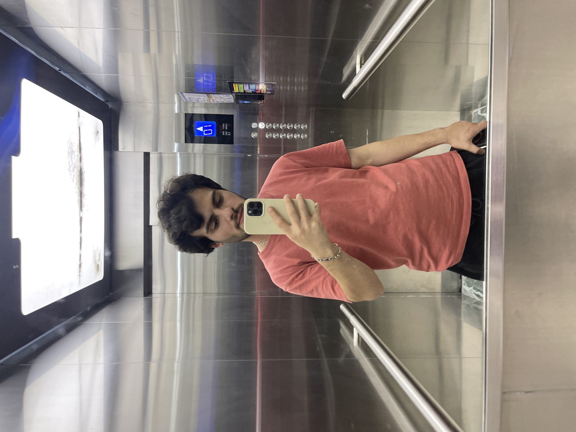 A young man with dark hair and a beard takes a selfie in an elevator in Tbilisi, Georgia. He is wearing a salmon-colored t-shirt and black shorts. The elevator has stainless steel walls and handrails. The elevator control panel shows floor number "01". The floor is reflected in the mirror.