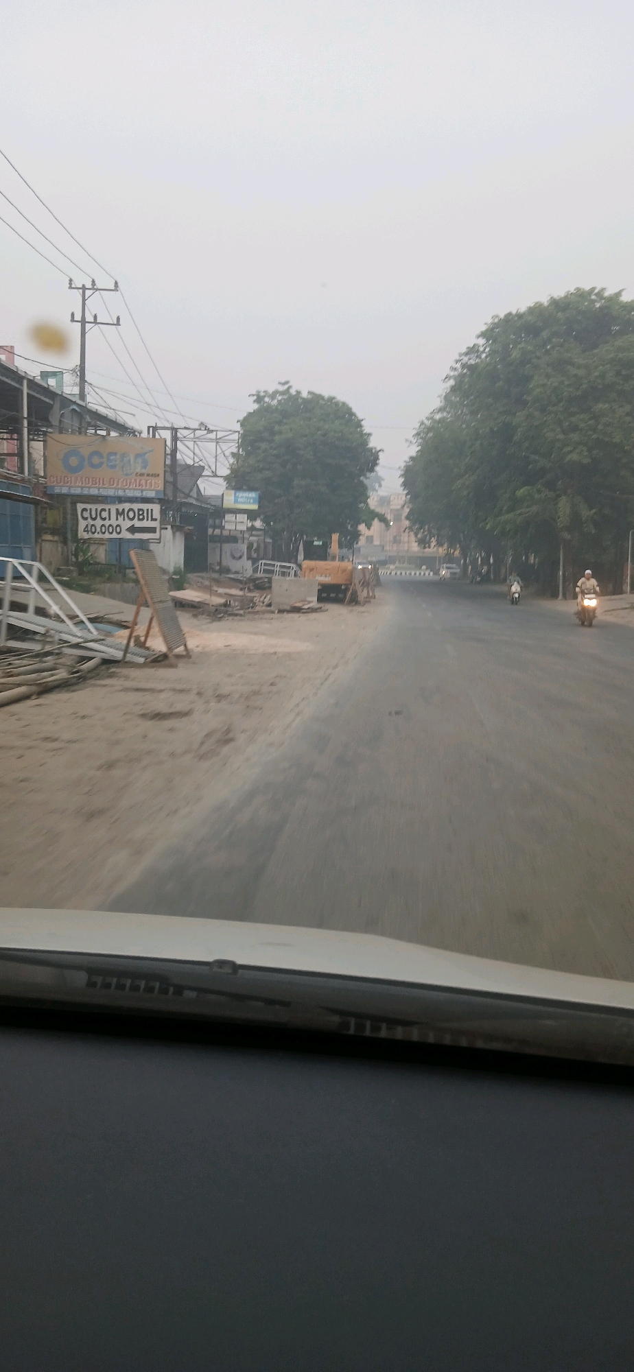The image shows a street scene in Samarinda, Indonesia. The weather is overcast. In the foreground, the dashboard and windshield of a vehicle are visible. The street is paved and appears to be undergoing construction or repair, with piles of debris, construction materials, and a yellow excavator on the side of the road. There are signs on the left side of the street, one of which reads "CUCI MOBIL OTOMATIS" (Automatic Car Wash) and another "CUCI MOBIL 40.000" (Car Wash 40,000). Trees line the sides of the road. Two motorcycles are visible on the road, one ridden by a person wearing a helmet. Electrical poles and wires are also present.