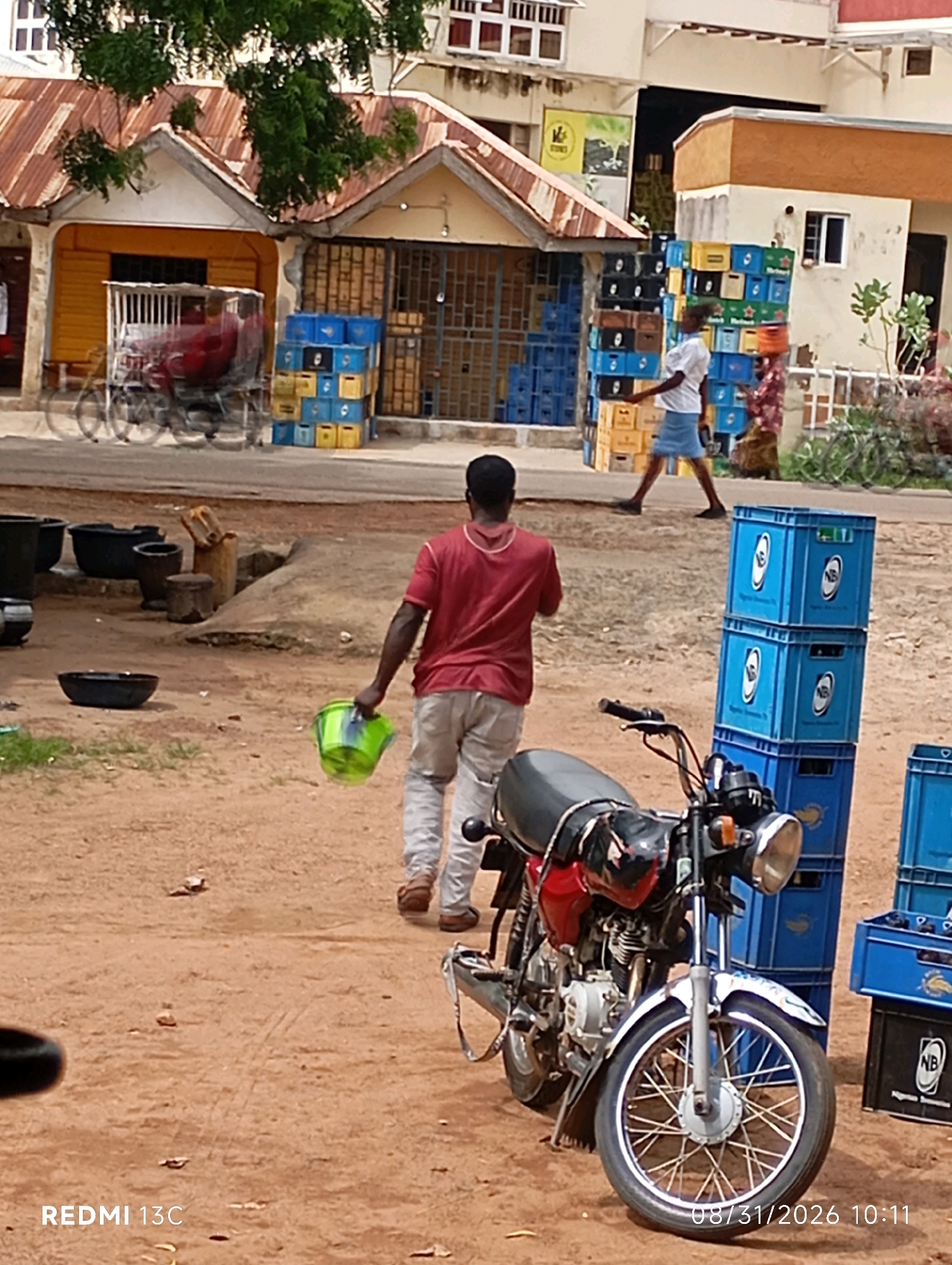 The image depicts an outdoor scene in what appears to be a market or commercial area. In the foreground, a red motorcycle is parked on a dirt ground. To the right of the motorcycle, stacks of blue plastic crates are visible. A man wearing a red shirt and light-colored pants is walking away from the viewer, carrying a green bucket in his right hand. Further in the background, a man in a white shirt and blue shorts walks past a large stack of crates. Buildings with corrugated metal roofs and storefronts line the street. Various large black pots and containers are placed on the ground in the middle ground. The environment suggests a casual, possibly commercial setting in a warm climate.