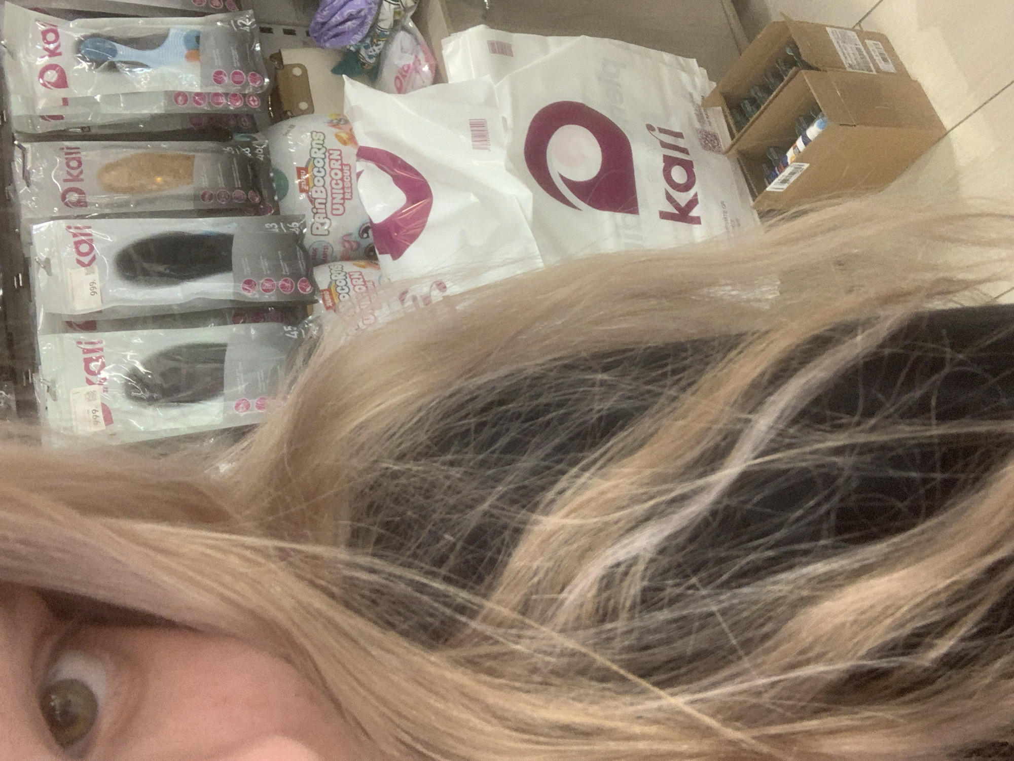 The image captures a retail scene from a first-person perspective, with a person's eye and blonde hair visible in the lower left foreground. The setting appears to be an indoor store or a retail storage area in Saint Petersburg, Russia.
The majority of the frame is filled with various products and packaging. On the left, several transparent packages containing what seem to be hair extensions or accessories are stacked on shelves. These packages feature the brand name "kari" and include numerical labels such as "999." (likely a price), "42," "43," "45," and "46." The contents of these packages are in different colors, including light blue, light brown, and dark brown/black.
In the middle and right background, several white plastic shopping bags are visible, each adorned with a prominent purple-pink logo and the "kari" brand name. Adjacent to these bags, colorful packaging for "RainBocorns UNICORN RESCUE" toys can be seen. Further right, a brown cardboard box containing various items is partially visible, indicating a stock or display area.
The overall impression is of a person browsing or observing merchandise in a retail environment.