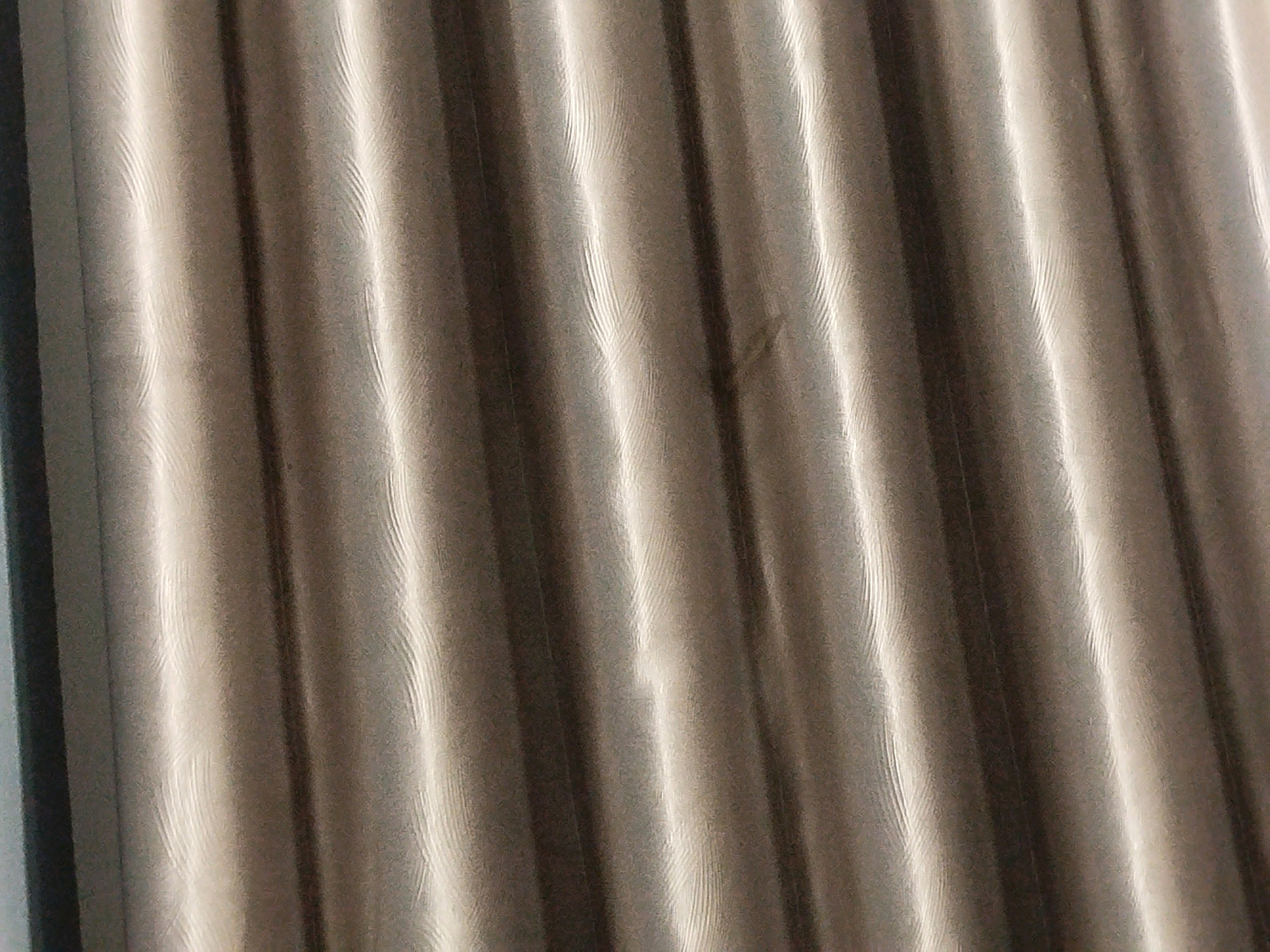 The image displays a close-up view of pleated curtains. The fabric is a neutral, taupe color with a subtle sheen that highlights the vertical folds. The lighting creates strong shadows and bright highlights, emphasizing the textured, wavy surface of the material. There are no people or other discernible objects visible in the frame. The scene is devoid of any context that would associate it with a specific location in Mojoagung, Indonesia, or any other place. The image is purely an abstract study of light and texture on fabric.