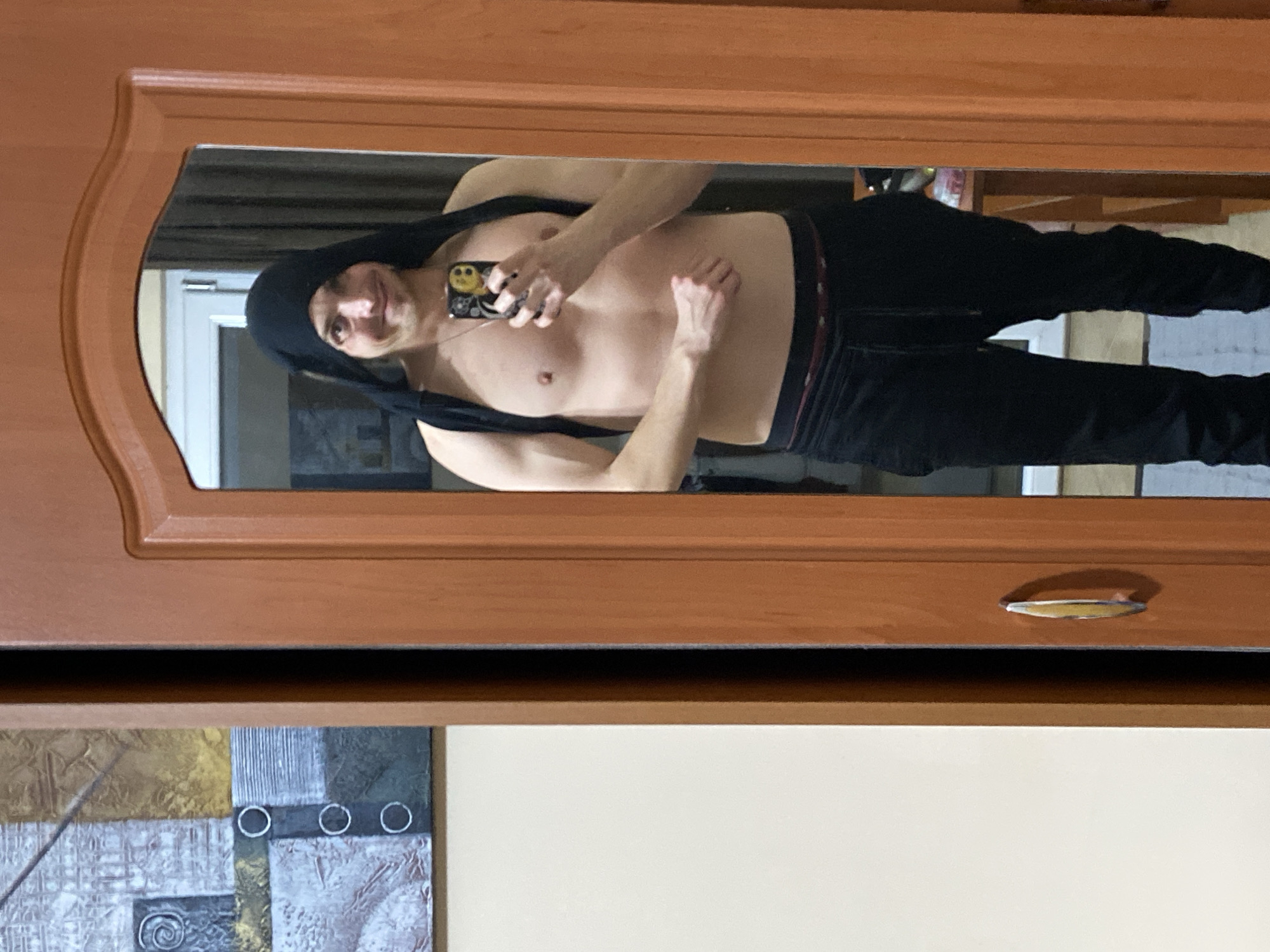 The image shows a reflection of a shirtless man in a mirror. He is wearing black pants and a black hood and is holding a phone with a yellow smiley face on the case. The mirror is framed with a brown wooden frame. Below the mirror is a drawer with a silver-colored handle. A section of a painting with geometric shapes and colors is visible on the wall below the drawer. The location is not discernible from the image.