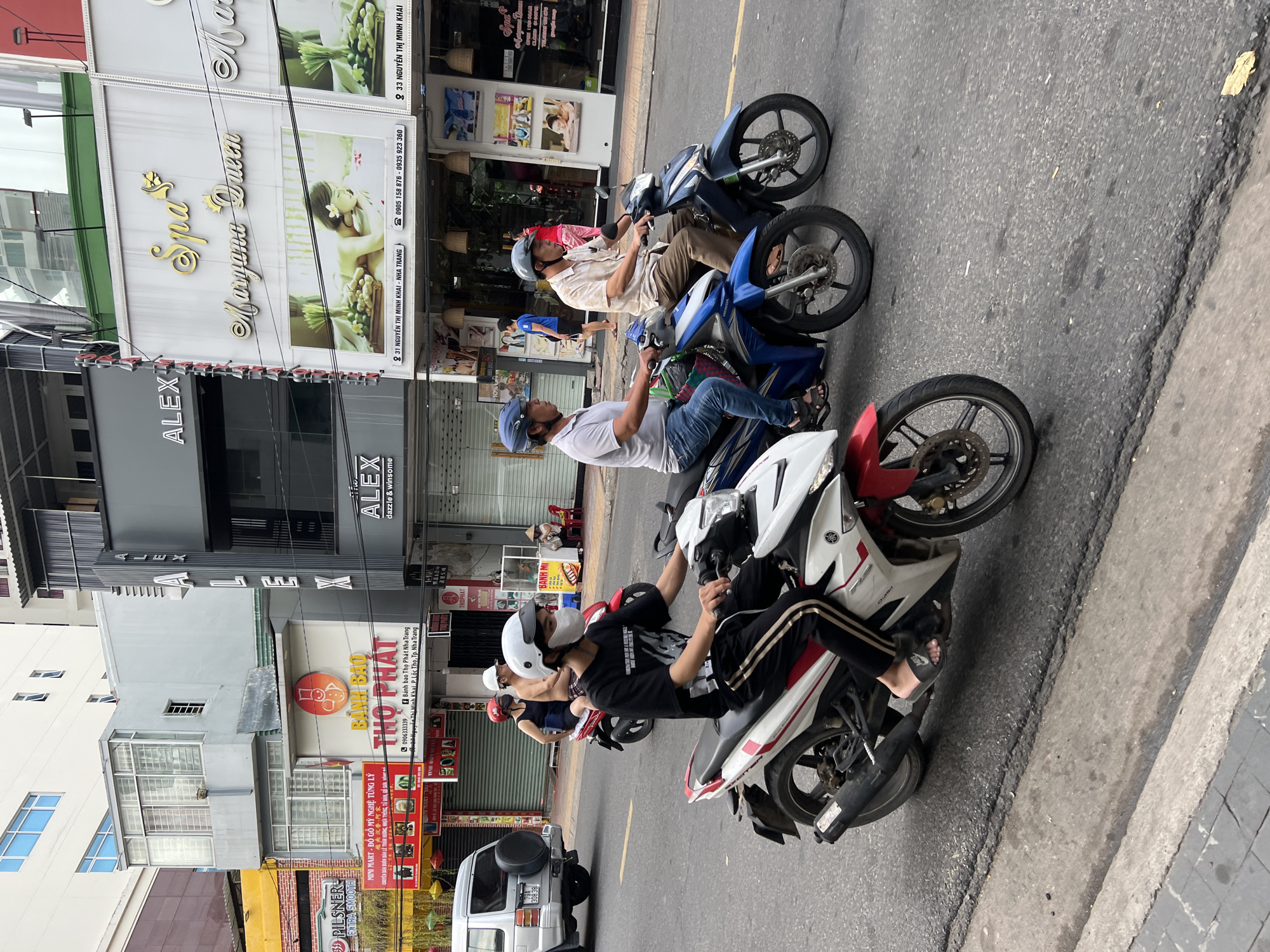 The image depicts a street scene in Nha Trang, Vietnam, with several motorbikes on a paved road. In the foreground, a young person wearing a white helmet and a white face mask rides a white and red motorbike. They are dressed in a black t-shirt and black pants with a white stripe. Behind them, a blue motorbike is ridden by a man in a white t-shirt and blue jeans, wearing a blue helmet. Another man in a light-colored patterned shirt and brown pants, also wearing a blue helmet, rides a dark blue motorbike to his right.
The background shows buildings lining the street. One building has signage for "ALEX" and "Spa Maryana Queen," with an image of a woman receiving a spa treatment. Another building has a sign with a red circle and a cartoon chef, advertising "Banh Bao Tho Phat Nha Trang." Further down the street, a white SUV is parked on the left side of the road. A few pedestrians are visible further back, one wearing a conical hat. The road surface is asphalt, with some wear and tear. The overall lighting suggests it is daytime.