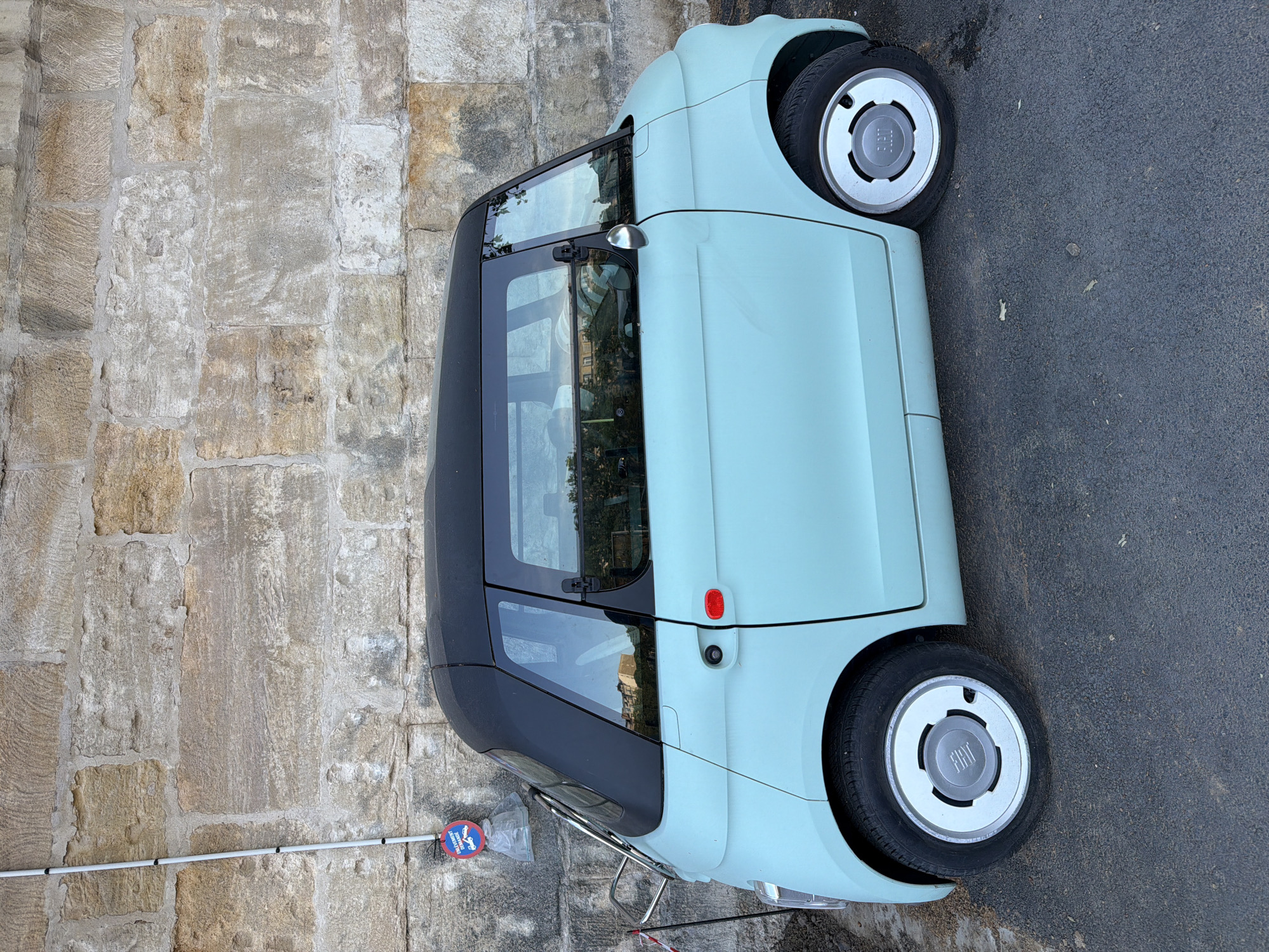 A pale blue Fiat 500e automobile with a black roof is parked on the street in front of a stone wall. The car has distinctive Fiat-branded wheel covers. A pole with a circular sign is affixed to the stone wall, likely indicating a parking restriction. The background includes urban elements suggesting a streetscape. Based on architectural style and the circular sign, the photograph was likely taken in Paris, France.