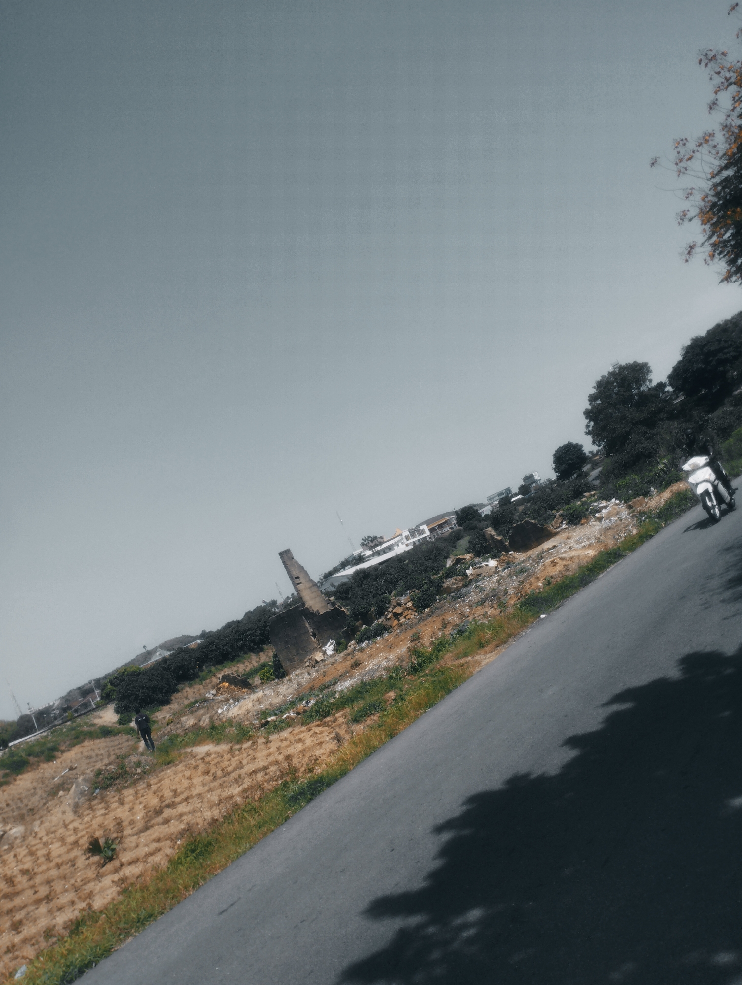 The image depicts a rural or semi-urban landscape with a road angled diagonally across the frame. A white motorcycle is traveling on the road, with a person on it. To the left of the road, there is a sloped terrain with dry, yellowish vegetation and scattered green patches. In the background, there are trees, some buildings, and what appears to be a partially ruined structure. The sky is overcast with a gray hue. The image is tilted, giving a sense of movement or an unconventional perspective. The overall scene suggests an outdoor environment in a location that is possibly undergoing development or is somewhat undeveloped.