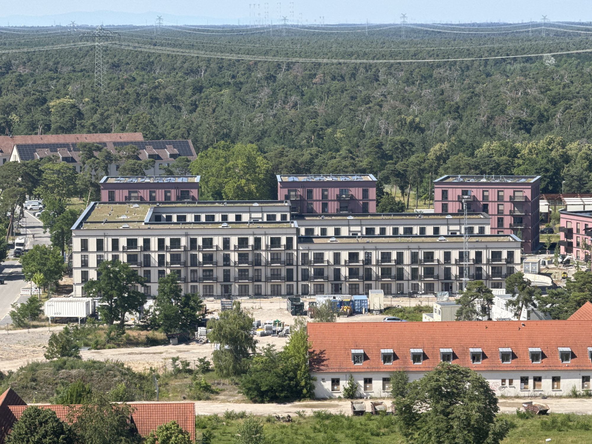 The image captures an aerial view of a section of Mannheim, Germany, likely a developing or redeveloping area on the outskirts of the city. In the foreground, a long, white building with a red tiled roof and multiple small dormer windows runs horizontally across the frame. To the right of this building, another red-roofed structure with multiple dormers is visible.
Dominating the middle ground are several modern apartment buildings. The largest of these is a multi-story, light-colored building with numerous balconies and dark-framed windows, appearing to be newly constructed or undergoing renovation. To its left and right are similar, though slightly smaller, buildings with reddish-pink facades.
The background is characterized by a dense forest stretching towards the horizon. Above the treeline, a network of high-voltage power lines supported by large transmission towers crisscrosses the sky, indicating the presence of significant electrical infrastructure.
Scattered throughout the scene are elements of construction and urban infrastructure, including what appear to be shipping containers, construction vehicles (such as a tractor and a forklift), piles of building materials, and paved roads with a few parked vehicles. The overall impression is one of a transition zone between a wooded natural environment and urban development.