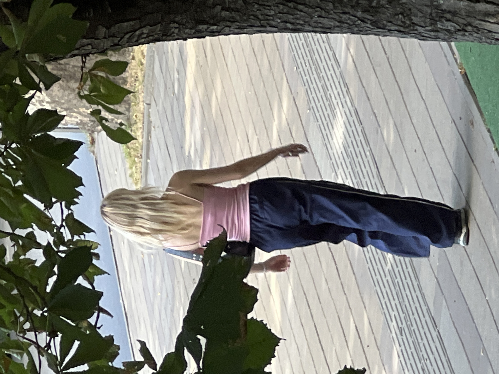 An unseen person walks away from the camera on a paved walkway next to a tree. The person has long, blonde hair and wears a pink tank top and dark blue pants with a white stripe. A dark bag is slung over their left shoulder. The walkway is made of light-colored planks with a textured strip running through it. Green grass is visible at the bottom right of the frame. The scene takes place outdoors in what appears to be a park or waterfront area, possibly in Krasnodar, Russia, given the context.