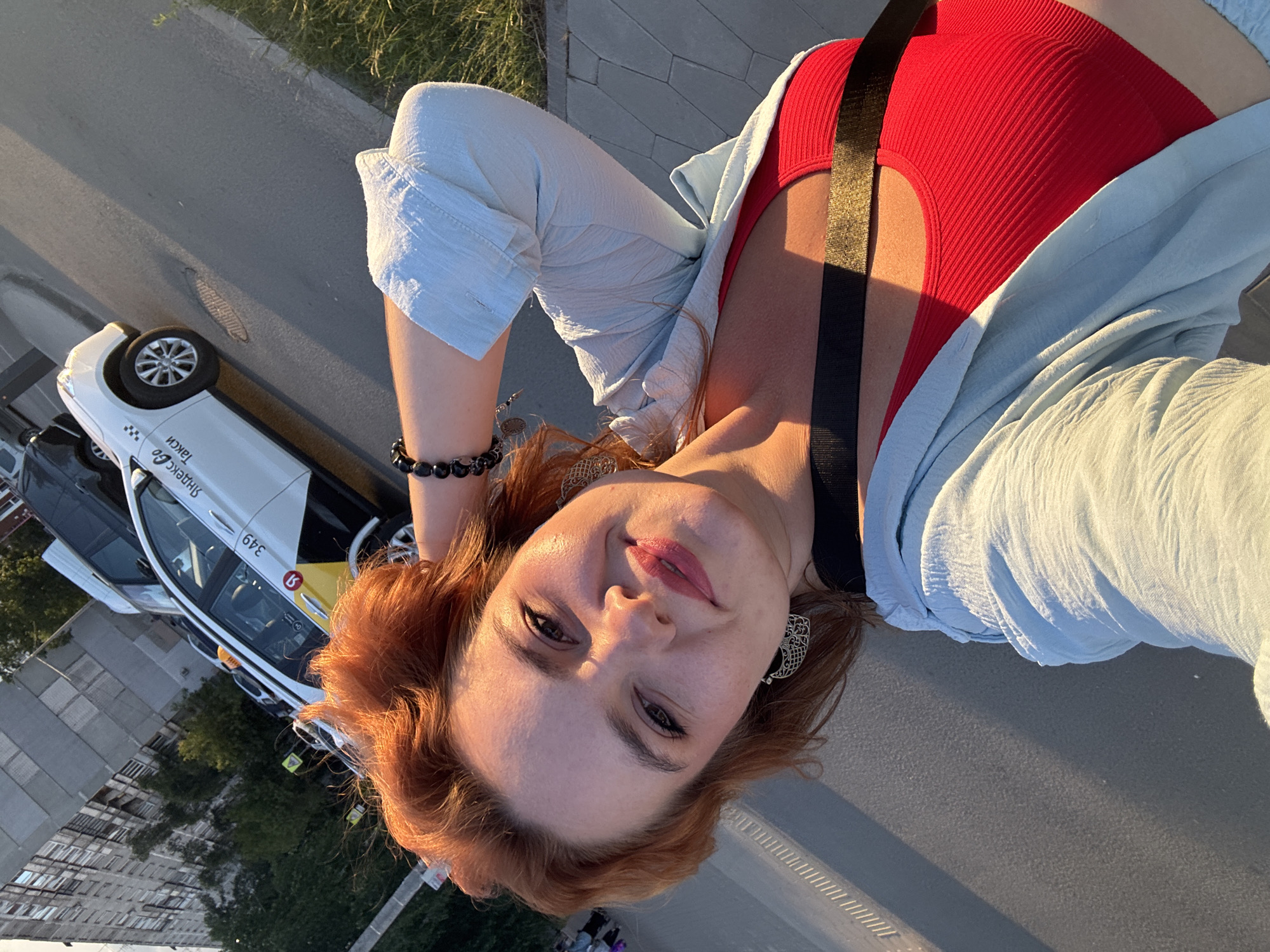 A selfie is taken from a slightly high angle outdoors during the day. A woman with red hair and makeup is in the foreground, smiling and touching her head. She is wearing a red tank top and a light blue unbuttoned shirt. A black strap is visible across her chest. Behind her, a white Yandex Go taxi with yellow accents and the number 349 on its door is parked on the side of a street. A tall, weathered apartment building with many windows is visible in the background, along with trees and a grassy area. The scene appears to be in an urban environment, likely Nizhny Novgorod, Russia, given the Yandex Go branding.
