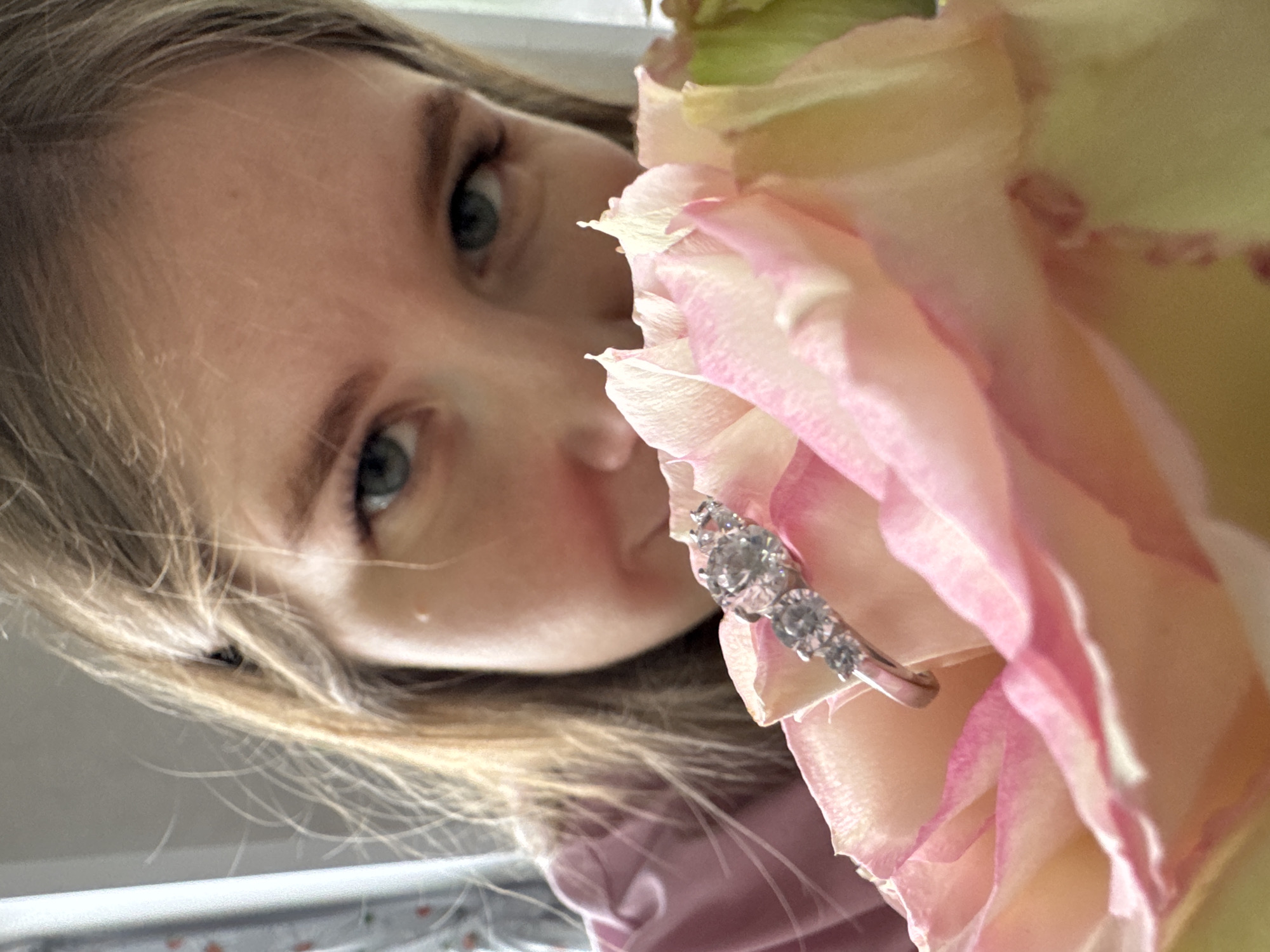 An indoor scene in Gornyi (Dvurechenskoe S/P), Russia, depicts a light-skinned female partially visible from the eyes up, peeking over a light pink and yellow rose. Her blue-green eyes are directed forward. She has light brown hair and is wearing a light pink garment. A silver-colored ring, adorned with five clear, round-cut stones of graduated size, rests atop the rose's petals in the foreground. The background is blurred, indicating an out-of-focus indoor environment with ambient light.