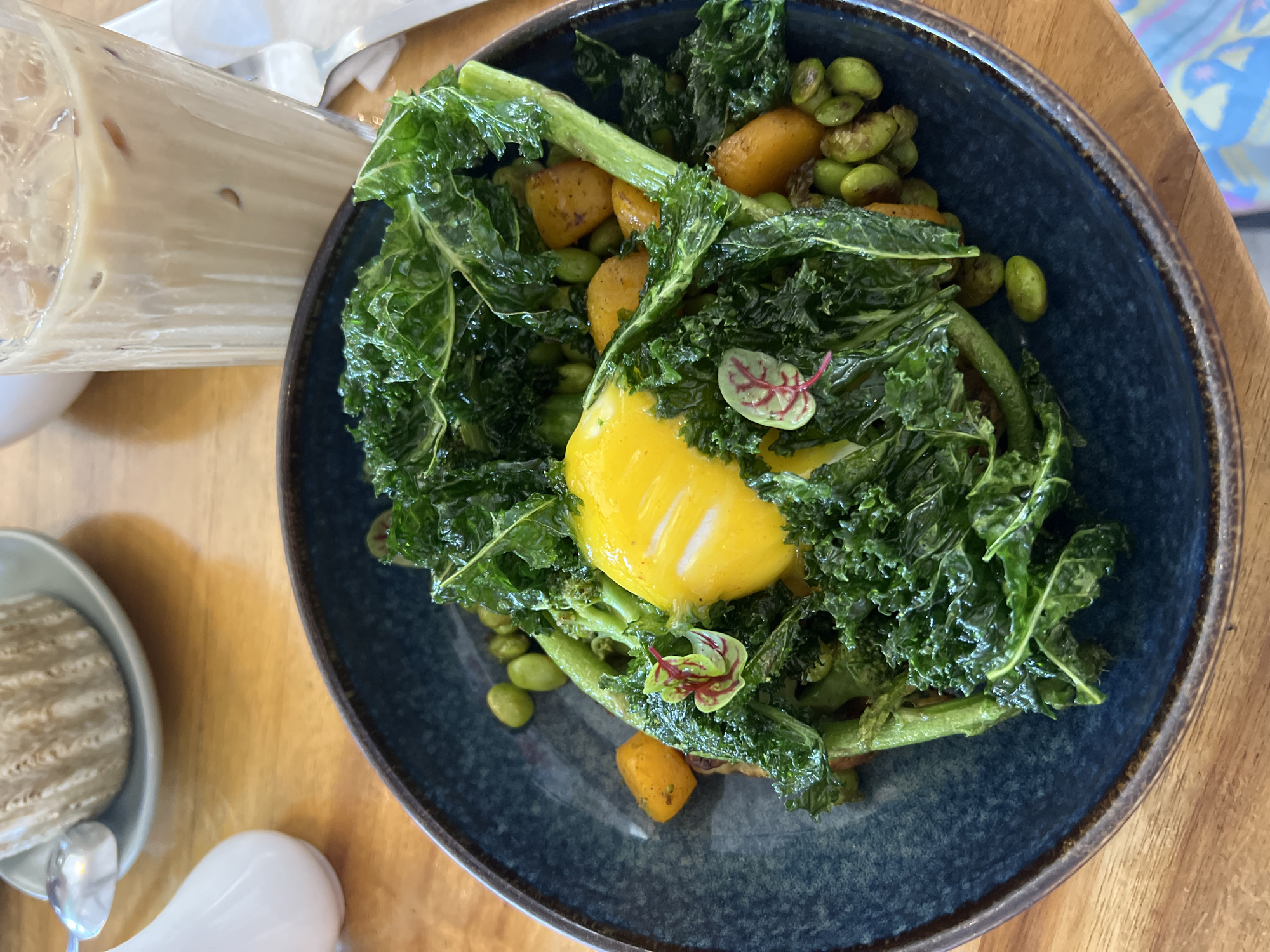 This close-up shot captures a healthy and colorful meal arranged on a dark blue ceramic plate with a textured, rustic rim, set upon a light brown wooden table. The central dish features a mound of vibrant green, leafy vegetables, likely kale, glistening with a light dressing. Nestled within the greens is a prominent bright yellow egg yolk, indicating a poached or soft-boiled egg, surrounded by green edamame beans and cubed orange vegetables, possibly roasted sweet potato or pumpkin. A slender green vegetable stalk or stem is also visible, along with a small, reddish-green microgreen used as a garnish.
To the left of the main plate, a tall glass filled with an iced, milky beverage, resembling iced coffee or chai, is partially visible. Further left, a small gray bowl contains a light-colored, textured snack, accompanied by a small silver spoon. A white, pear-shaped dispenser or container is also seen at the bottom left. The overall arrangement suggests a fresh and appealing dining experience, typical of a cafe setting. The presence of an iced drink might indicate a warm climate, aligning with the North Kuta, Indonesia location. The scene portrays a moment before or during a meal, likely brunch or lunch, given the components of the dish. No individuals or discernible text are present in the image.