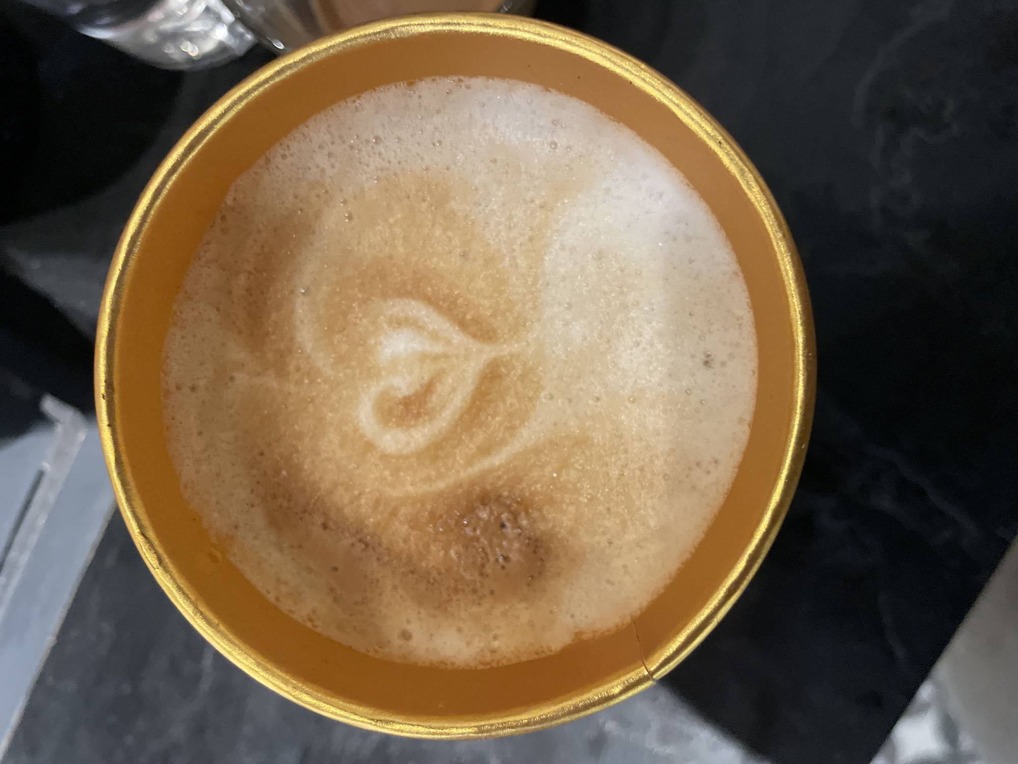 The image shows an overhead view of a cup of coffee, possibly a cappuccino or latte, with a heart shape formed in the foam. The cup is gold-colored and appears to be sitting on a dark surface, possibly a table. Parts of another cup/glass are visible in the top left corner. The overall lighting is somewhat diffused, creating soft shadows. There are no people visible in the shot. The photograph’s location in Veliko Tarnovo, Bulgaria is unknown, or not evidenced by any clues within the image.