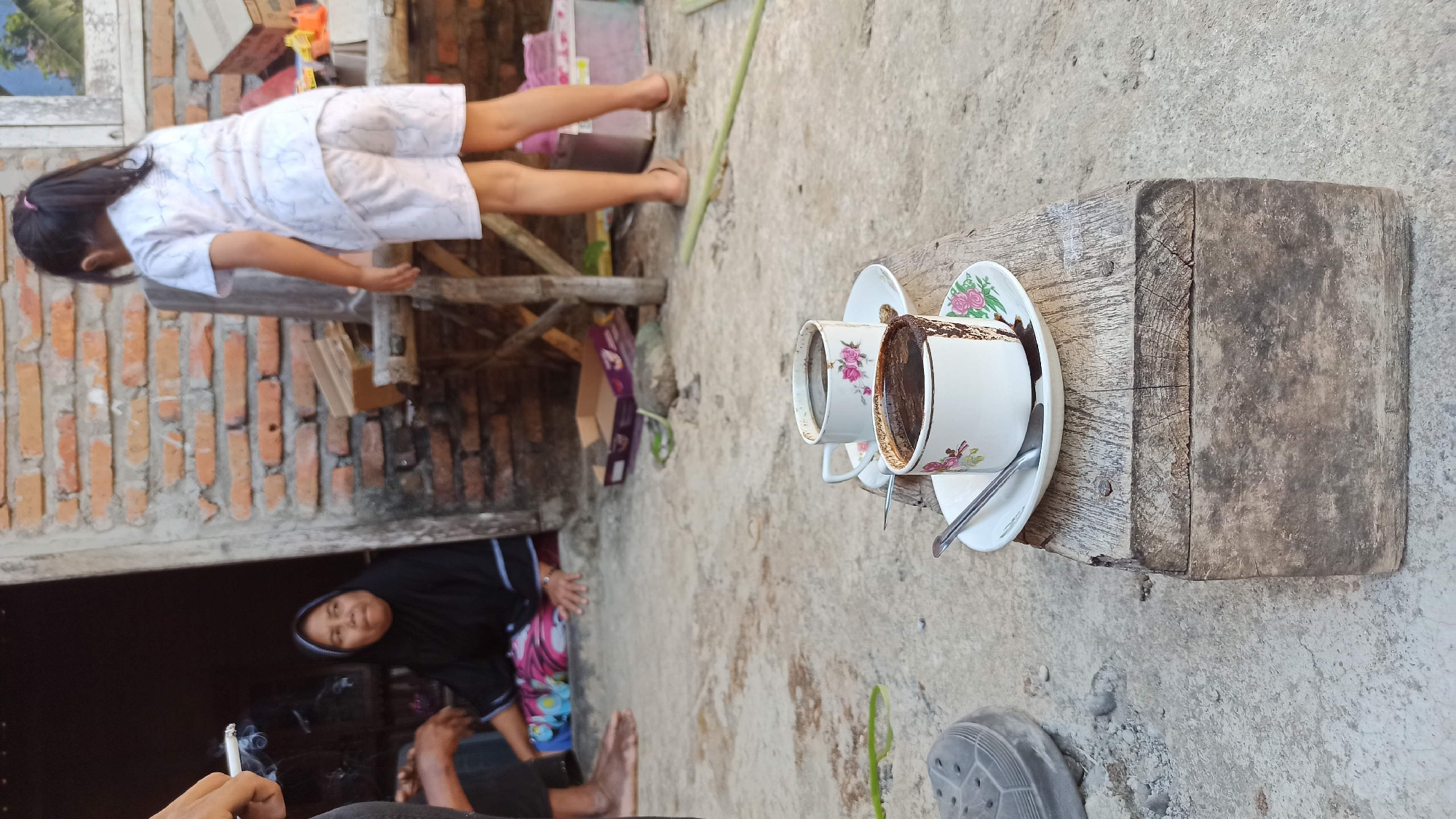The image depicts an outdoor scene in Gegeran, Indonesia, where two cups of coffee are placed on a rustic wooden block. In the background, a woman wearing a black hijab and a colorful sarong is seated, looking to the left. To her right, a young girl in a white t-shirt and shorts stands facing away from the camera. The environment appears to be an unpaved area with a brick wall and various objects scattered around, suggesting a residential or communal space. The lighting indicates it is daytime, likely late afternoon due to the warm tones.