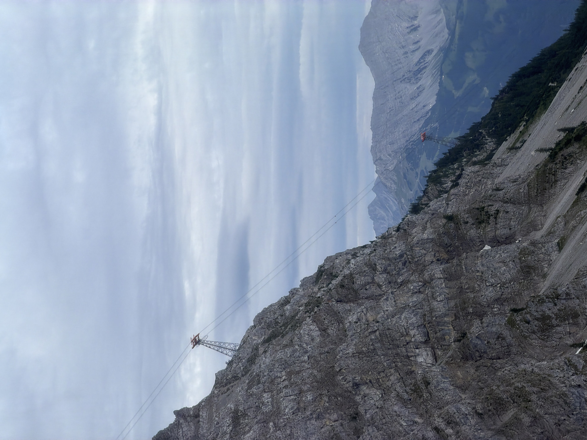 The image shows a steep, rocky mountain slope with a cable car support tower and cables in the foreground. The mountain is covered in gray, weathered rock with some sparse vegetation. In the distance, more mountains are visible, shrouded in mist and clouds. The sky is overcast with a pale, grayish hue. A red cable car support tower is visible on the slope, with cables stretching upwards and across the frame. This scene is consistent with the Zugspitze mountain, the highest peak in Germany, located near the town of Grainau. There are no people visible in the image.