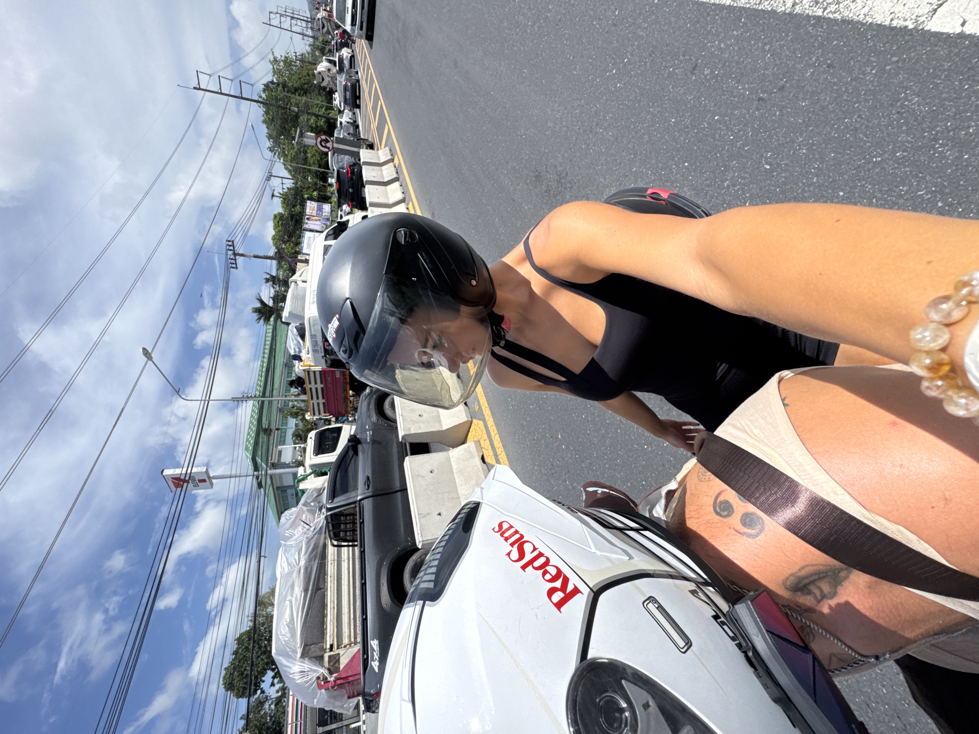 A woman wearing a black helmet and black tank top sits on a motorcycle in Wichit, Thailand. She appears to be in traffic, with a line of cars visible in the background along the side of the road. The motorcycle has the word "Redsuns" written on it. The woman has a tattoo on her right thigh and a beaded bracelet on her right wrist. A 7-Eleven store is visible in the background.