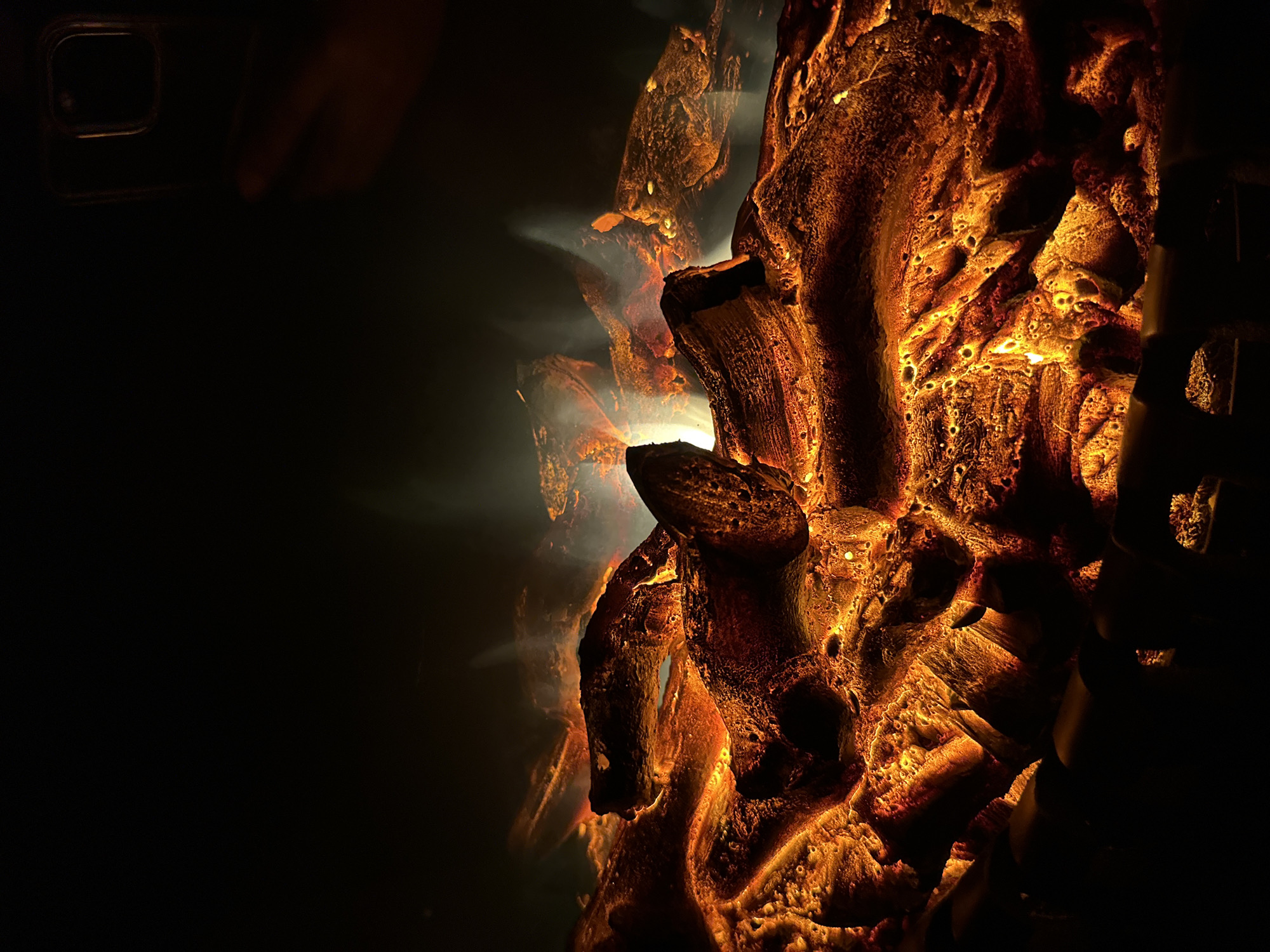 The image depicts a close-up, low-angle shot of artificial logs and embers in a fireplace, casting a warm, orange glow against a dark background. The logs appear textured and irregular, with some emitting faint wisps of smoke or steam. The embers beneath are a vibrant mix of yellow and orange, creating a dynamic and lively effect. In the upper right corner, a hand is visible, holding what appears to be a smartphone, suggesting someone is taking a photograph or video of the fireplace. The overall ambiance is cozy and inviting. The location is listed as Unknown, Unknown, indicating that specific geographical or urban details are not available.