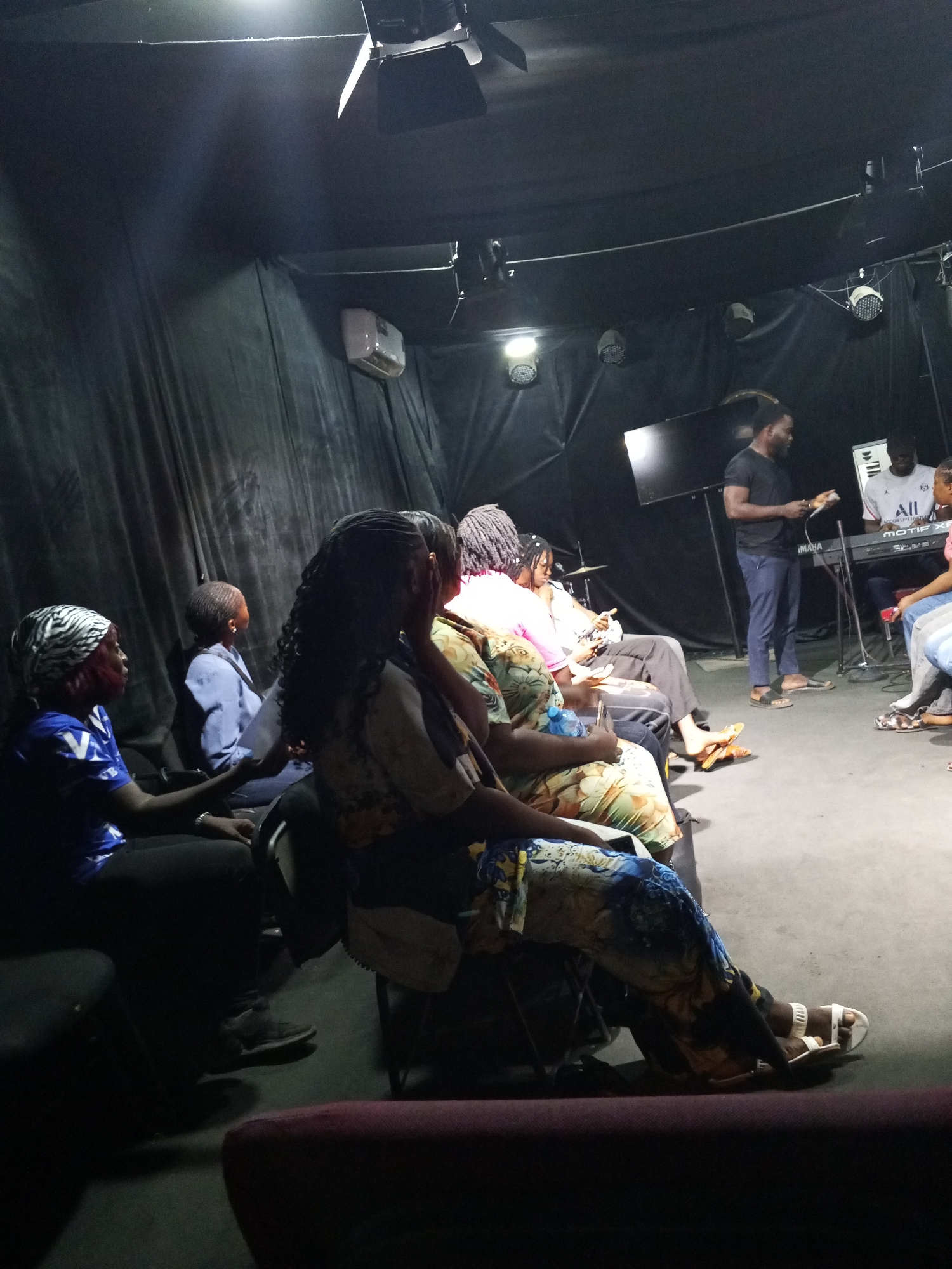 The image depicts a dimly lit interior, possibly a small performance space or theater, located in Abuja, Nigeria. Several people are seated in rows of chairs facing a man standing and holding papers or a small book. Behind him, a keyboard and monitor are visible, along with other people possibly involved in a performance or presentation. The environment has black curtains hanging and stage lighting equipment mounted on the ceiling. The individuals in the foreground are wearing casual attire, and the overall atmosphere appears to be intimate and informal.