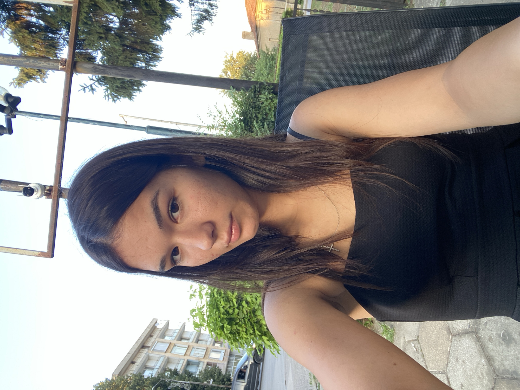 A young woman with long, dark brown hair is pictured sitting outdoors in Sevlievo, Bulgaria. She is wearing a black, strapless dress and a silver necklace with a cross pendant. Her right arm is visible, resting on the armrest of a dark grey chair. In the background, a building with multiple floors and balconies, lush green foliage, and a street with a car can be seen. The sky is clear and bright. The overall scene suggests a casual outdoor setting, possibly a cafe or park in the city of Sevlievo.