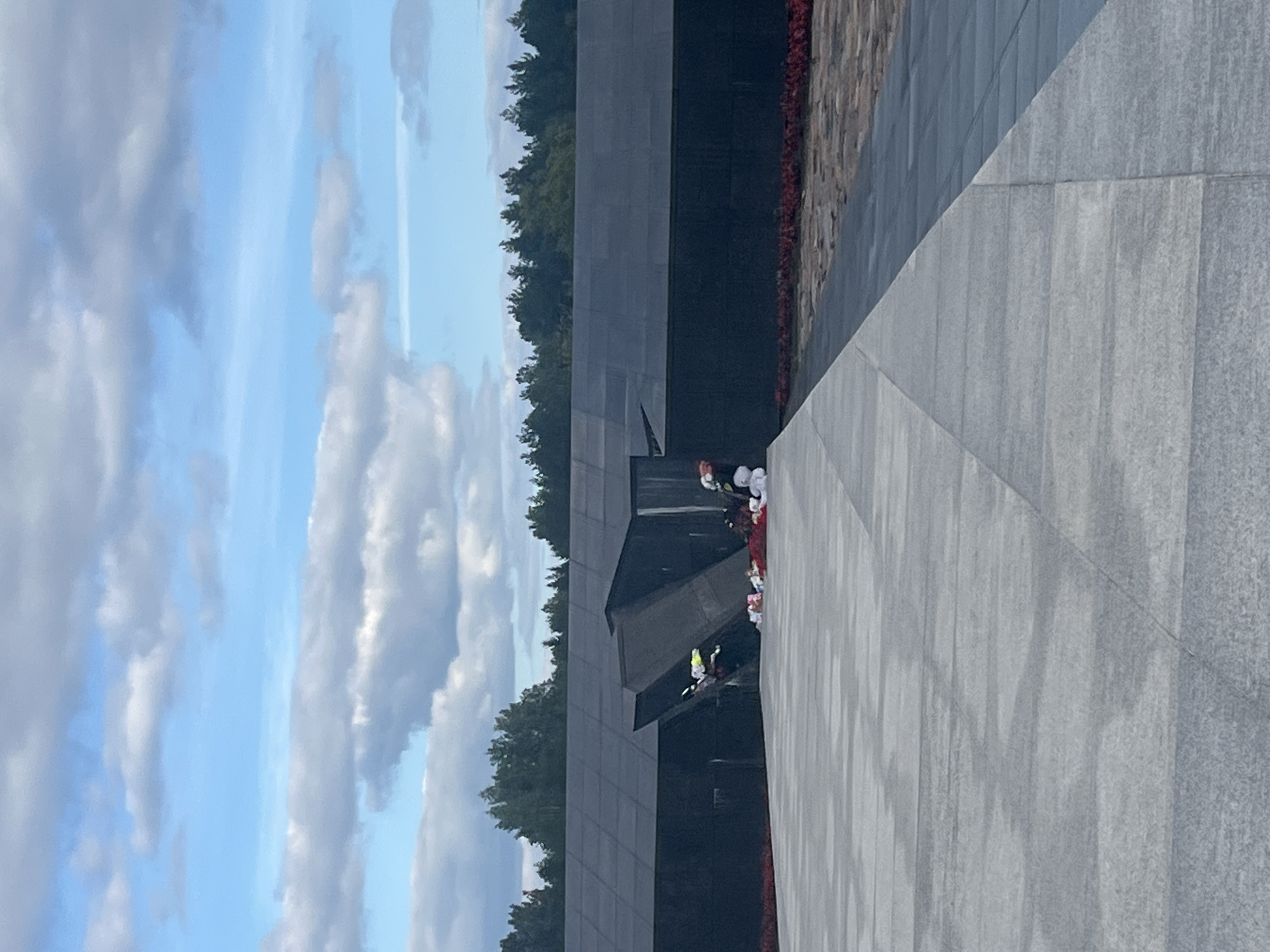 The image captures a somber monument with stark architectural elements against a cloudy sky. A large, dark, angular structure, possibly a memorial, dominates the background, with a large opening or fissure. Several bouquets of flowers, predominantly white and red, are placed at the base of this structure and on a paved area in the foreground. The ground is paved with large, light-colored stone slabs, forming a geometric pattern. A line of red flowers or ground cover runs along the base of the dark structure. Trees form a dark green line along the horizon behind the monument. The overall impression is one of solemnity and remembrance. The location is the Khatyn Memorial Complex, situated within the territory of Kamenski Sel'ski Savet, Belarus, commemorating victims of Nazi atrocities during World War II.