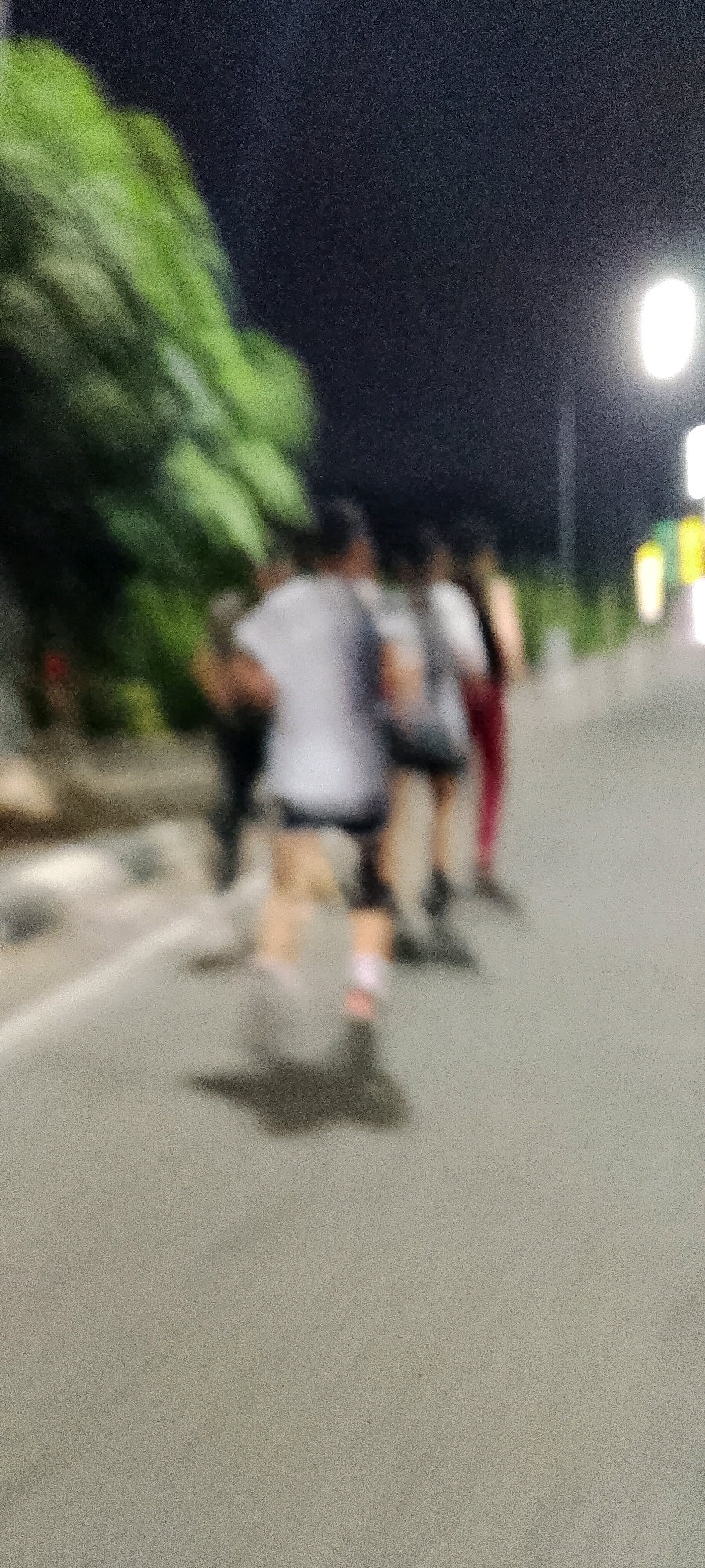 The image shows a blurry, nighttime scene with several people running on a paved road. To the left, there are trees with green foliage. In the background, there are streetlights casting a glow. The people appear to be dressed in athletic wear, with one person in the foreground wearing a white t-shirt and dark shorts, and another in the middle ground wearing a white shirt and dark pants. The overall impression is of an outdoor running activity in an urban or suburban environment in Gunung Putri, Indonesia.