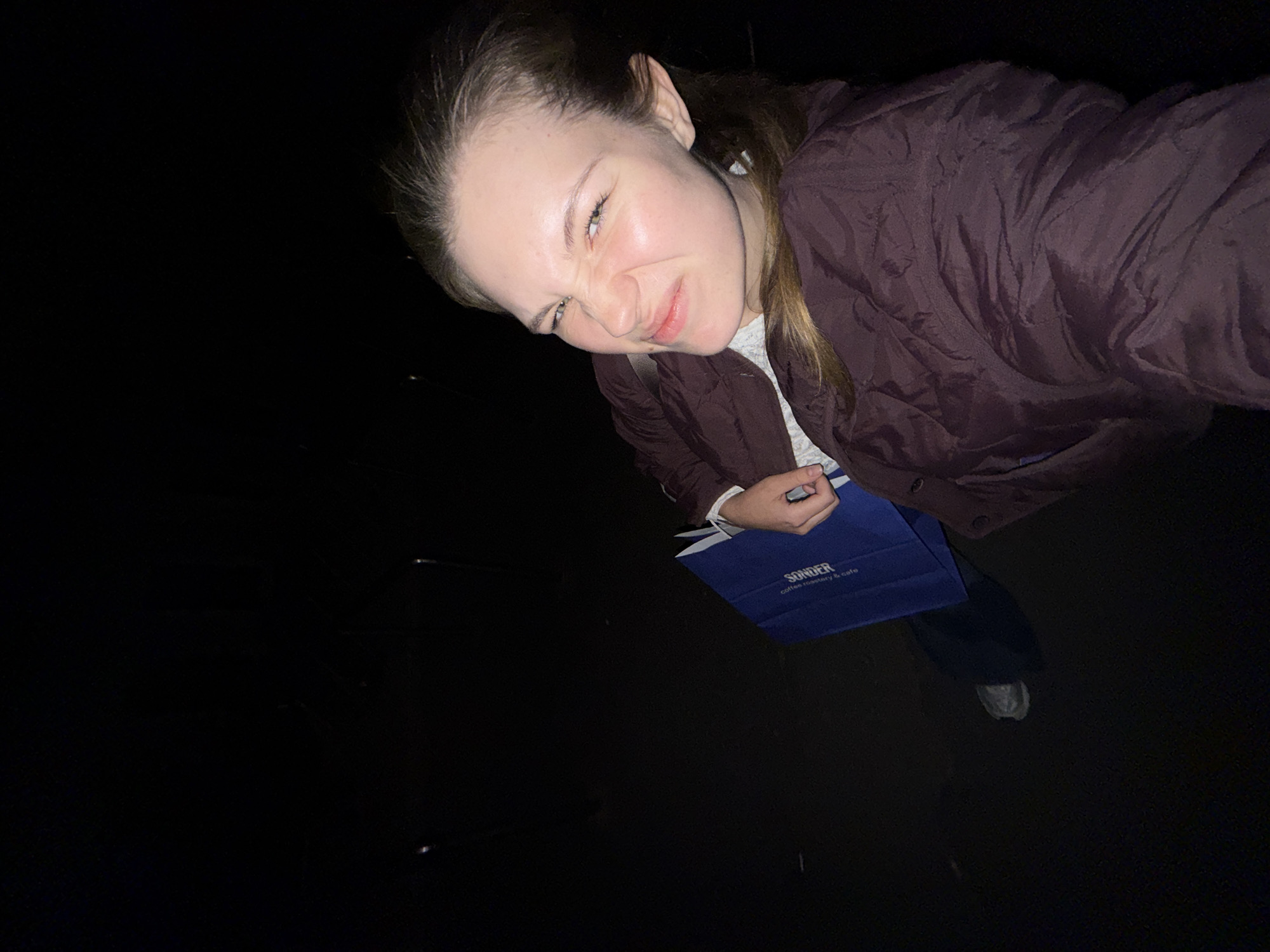 A woman stands outdoors in Belgrade, Serbia at night. The view is from a high angle, as if the photo was taken in a selfie style. The woman is wearing a puffy, brown jacket and jeans, and she holds a blue bag that has "SONDER coffee roastery & cafe" printed on it. The woman is making a face, scrunching up her nose and squinting her eyes. The background is dark.