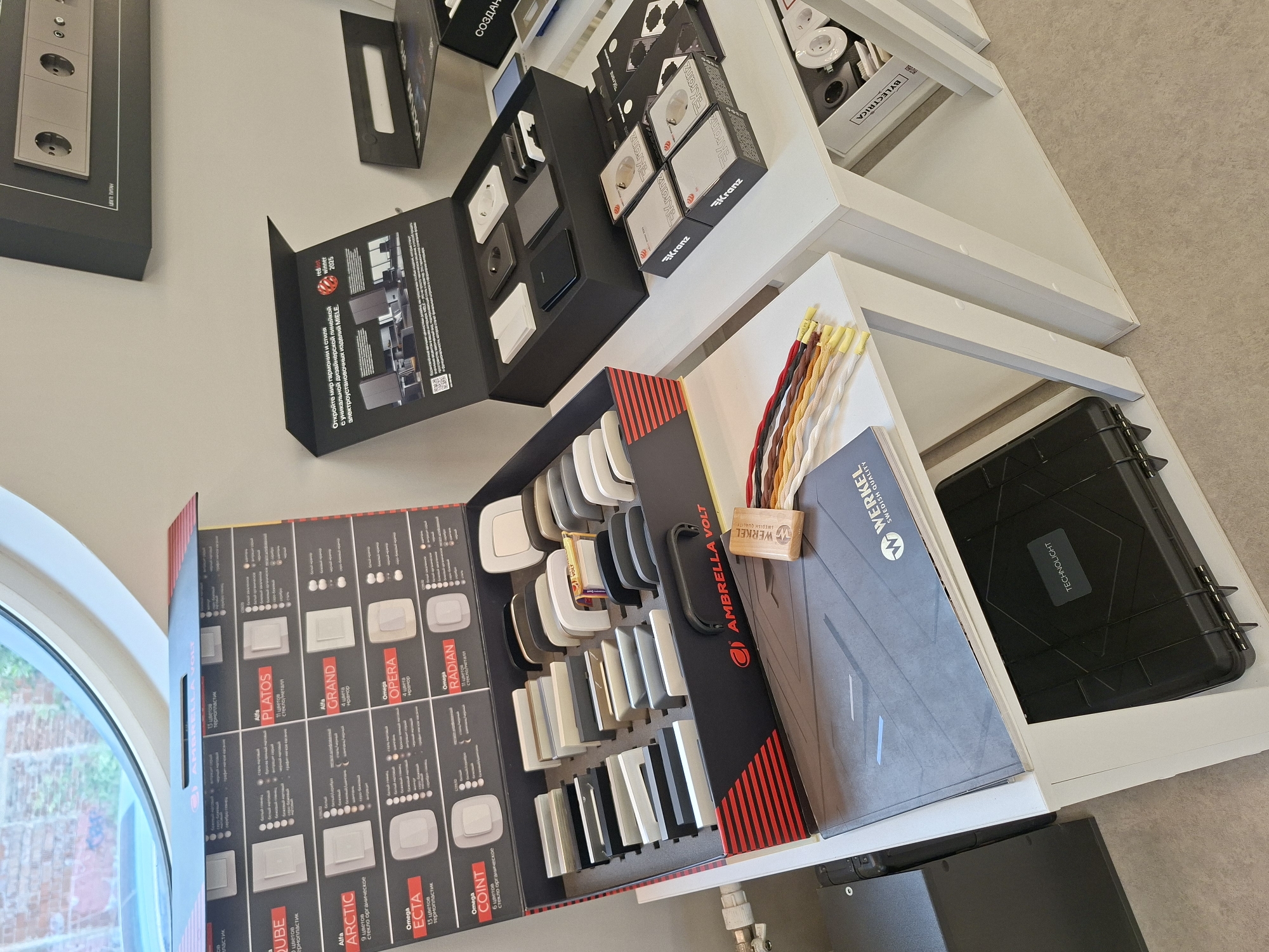 The image depicts an interior display of electrical fittings and samples, likely in a showroom or retail store. The items are organized on white shelving units.
On the top shelf, to the left, is an open box showcasing various color and material samples of electrical plates, with branding visible as "AMBRELLA VOLT". Next to it, to the right, is a black box display with several individual electrical sockets and switches, some of which have "MIELE" branding. Further to the right on the top shelf, a framed image of electrical sockets is visible.
On the middle shelf, a collection of smaller boxes, some with the "Kranz" logo, are arranged. There are also open boxes with electrical fittings and samples visible. A folder with "WERKEL SWEDISH QUALITY" branding lies on this shelf. A wooden block with colorful cords is also present.
On the bottom shelf, a black industrial-style case with the label "TECHNOLIGHT" is placed. To the right, partially visible, is another shelf containing more electrical components.
The environment appears to be well-lit, with neutral-colored walls and flooring. The overall impression is of a professional display of electrical products. There is no indication of the city of Izhevsk, Russia in the image itself.