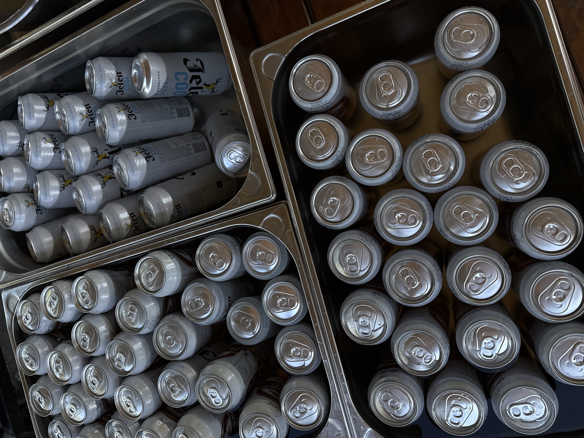 The image shows a top-down view of three rectangular metal trays filled with cans of beer. The tray on the upper left contains cans of "Jelen Cool" beer, stacked in a semi-organized fashion. The cans have a white and silver color scheme with black lettering and a stylized deer emblem. The other two trays are filled with cans of a different brand of beer, with silver tops and bodies, and a floral design at the rim of the can. The cans in the other trays are more uniformly arranged. The trays appear to be resting on a dark wooden surface.
Based on the brand of beer identified in the upper left tray, "Jelen Cool", it's possible this image was taken in Serbia, as "Jelen Pivo" is a popular Serbian beer. Further, given that Cukarica is the municipality location, it's plausible the image was taken at a restaurant, bar, or event venue within Cukarica.