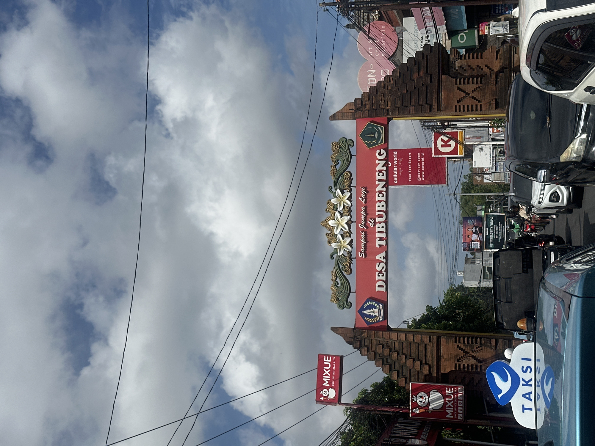 The image captures a street scene in North Kuta, Indonesia, featuring a prominent brick archway with ornate carvings and floral motifs. A red banner across the arch reads "Sampai Jumpa Lagi di DESA TIBUBENENG" with the text "cellular world" below it. A logo for "MIXUE" (ICE CREAM & TEA) is visible on a red sign to the left of the arch. On the right, a "K" logo within a red circle is displayed. The street is congested with traffic, including cars, motorcycles, and a blue taxi in the foreground with a "TAKSI" sign on its roof. Buildings and various advertisements line the sides of the road. The sky above is partly cloudy with a bright blue sky peeking through.