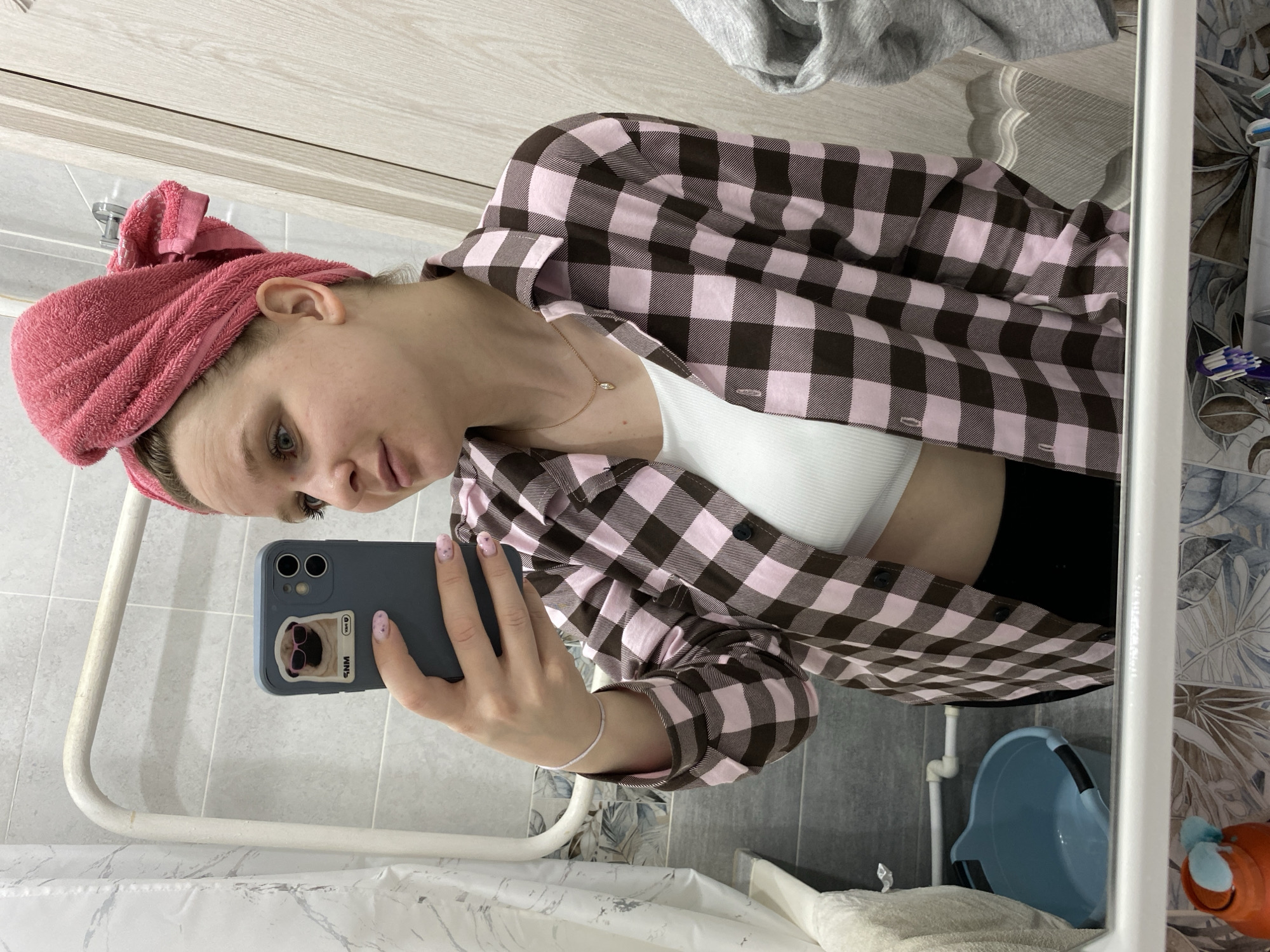 A woman in a bathroom takes a mirror selfie. She wears a pink towel on her head, a white tank top, and a pink and brown checkered shirt. She holds a blue smartphone with a pug design. The bathroom has gray and white marble-patterned walls and tiles. A small blue tub and orange object are visible in the lower right corner of the mirror.