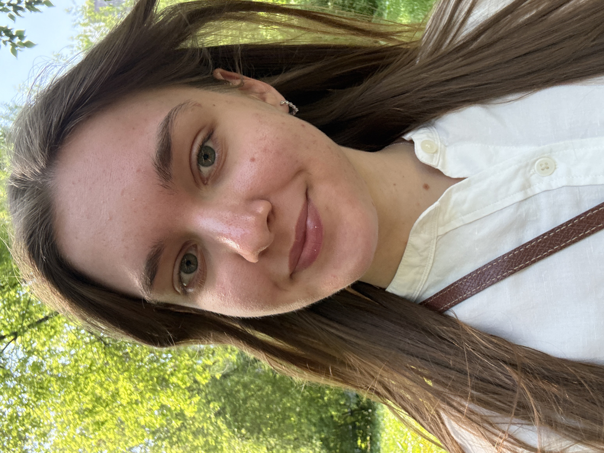 A selfie image shows a young woman in an outdoor setting. The woman has fair skin, brown hair, and green eyes. She is wearing a white shirt and a small earring. A brown strap is also visible, possibly from a bag or purse. The background shows greenery, suggesting trees and foliage. There is insufficient detail to determine the specific location in Perm, Russia.