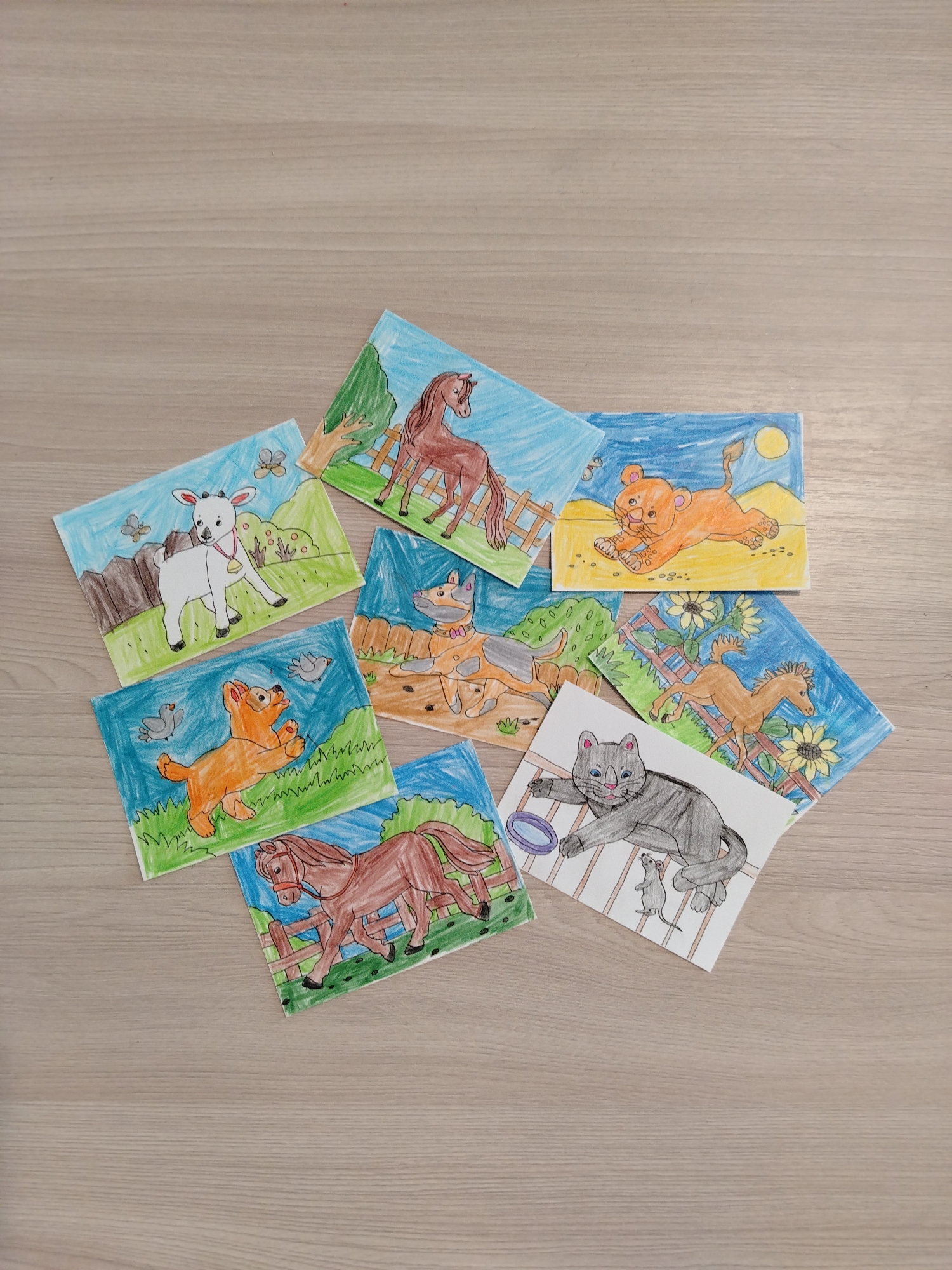The image shows a collection of eight hand-drawn, colorful animal pictures scattered on a light-colored wooden surface. The drawings feature a variety of animals including a goat, horse, lion cub, dog, and cat. Each drawing has a background setting, ranging from fields and fences to sunflower patches and night skies. The pictures are not aligned in any particular order and are loosely overlapping.
Location: The city of Chaikovsky, Russia, cannot be determined from the image.
