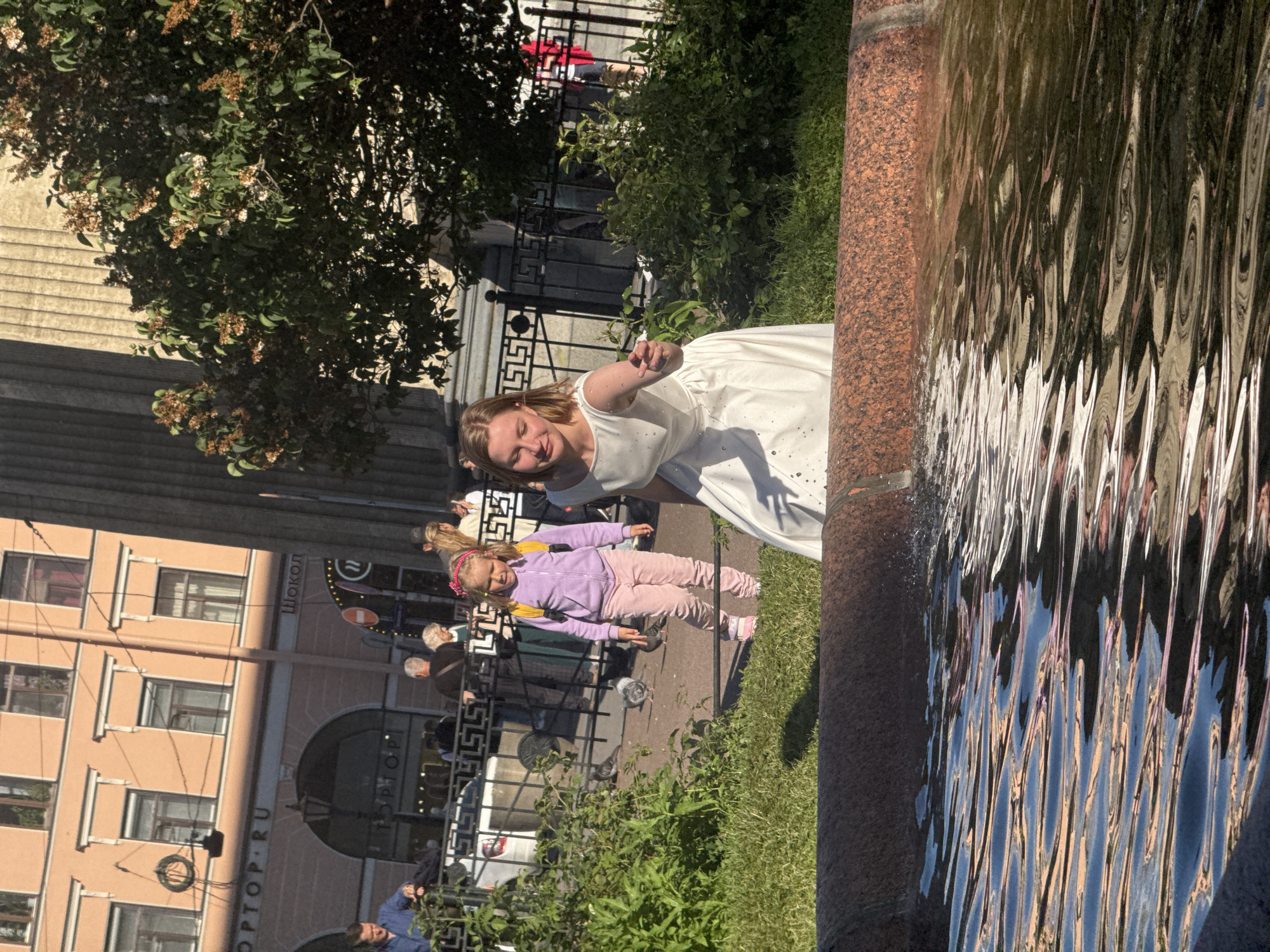 A young girl in a white dress stands near a fountain, her hand outstretched. Beside her, a younger girl in a lavender hoodie and pink pants walks. The scene appears to be outdoors on a sunny day, with lush green grass and trees. Behind them, a multi-story building with a pale peach facade is visible, featuring numerous windows and balconies. There's also a black metal fence with decorative elements. Visible text on the building includes "ШОКОЛ" and "TOP.RU". The water in the fountain reflects the surrounding environment, creating a distorted, rippling effect. The overall impression is a bright, leisurely moment captured in an urban setting.