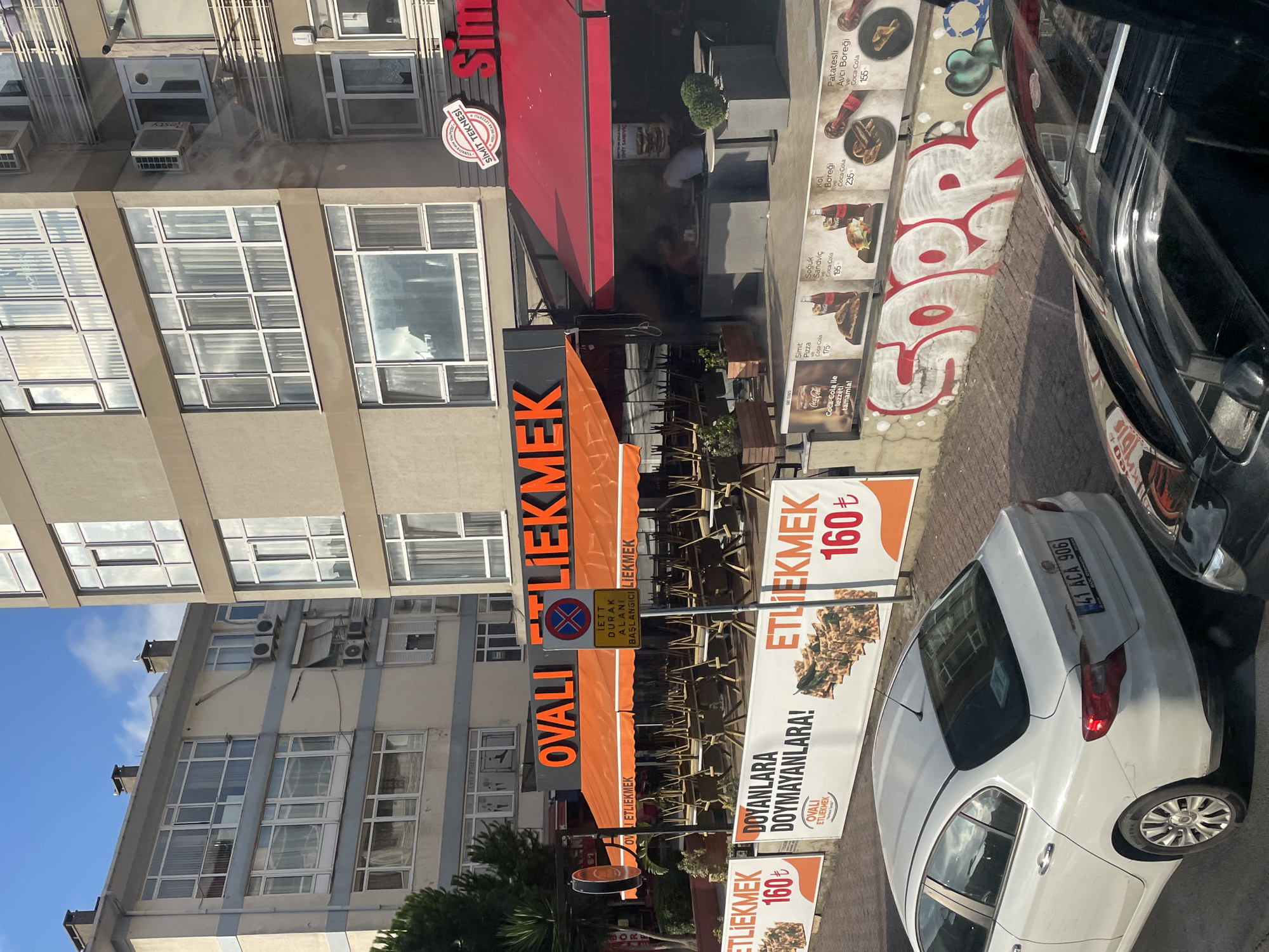 The image shows a street scene in Kadıköy, Turkey, on a sunny day. In the foreground, a white car with license plate "41 ACA 906" is visible. Behind it, a black car's front end is partially visible. The middle ground features a restaurant with an orange awning and large orange lettering that reads "OVALI ETLİEKMEK". Chairs are stacked neatly under the awning, suggesting the restaurant may be closed or preparing to open. A traffic sign prohibiting parking is posted in front of the restaurant. Further back, a taller building with multiple windows is visible. The background shows more buildings and a clear blue sky with some clouds. A banner advertising "ETLİEKMEK 160₺" is displayed on the left side of the image. Another banner with food images and prices is on the right side, displaying "Simit Pizza," "Soğuk Sandviç," "Kol Böreği," and "Patatesli Avcı Böreği." Graffiti, including the word "SORRY" in large red and white letters, is present on a wall below the banners. The overall impression is of a commercial street in a Turkish city.
