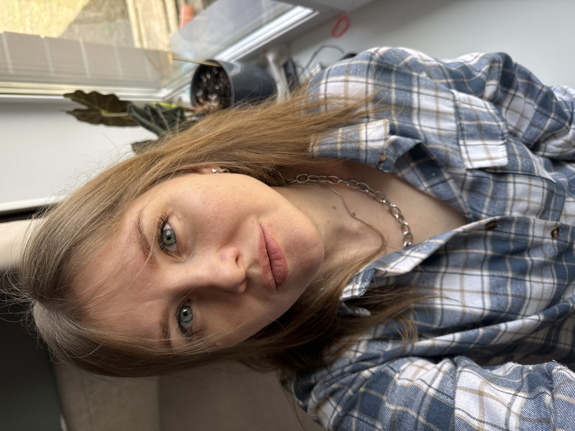 The image, rotated 90 degrees clockwise, displays a close-up portrait of a young woman, likely in her late teens or early twenties. She has light brown or ash blonde hair and striking blue-green eyes, which gaze directly at the viewer with a neutral to softly serene expression. Her lips are a natural pink hue, suggesting a subtle, closed-mouth smile. A small, silver heart-shaped stud earring is visible in her left earlobe.
She is dressed in a casual plaid flannel shirt, featuring a pattern of blue, white, and tan or light brown squares. A silver-colored chunky chain necklace is prominent around her neck, resting against her collarbone.
The background, softly blurred, indicates an indoor setting, likely near a window, given the natural light and faint reflections. A dark-leaved potted plant is visible behind her, along with another black pot, suggesting a domestic or well-maintained environment. The lighting suggests the photo was taken during the daytime. There is no visible text in the image. The location context provided is Gornyi (Dvurechenskoe S/P), Russia.