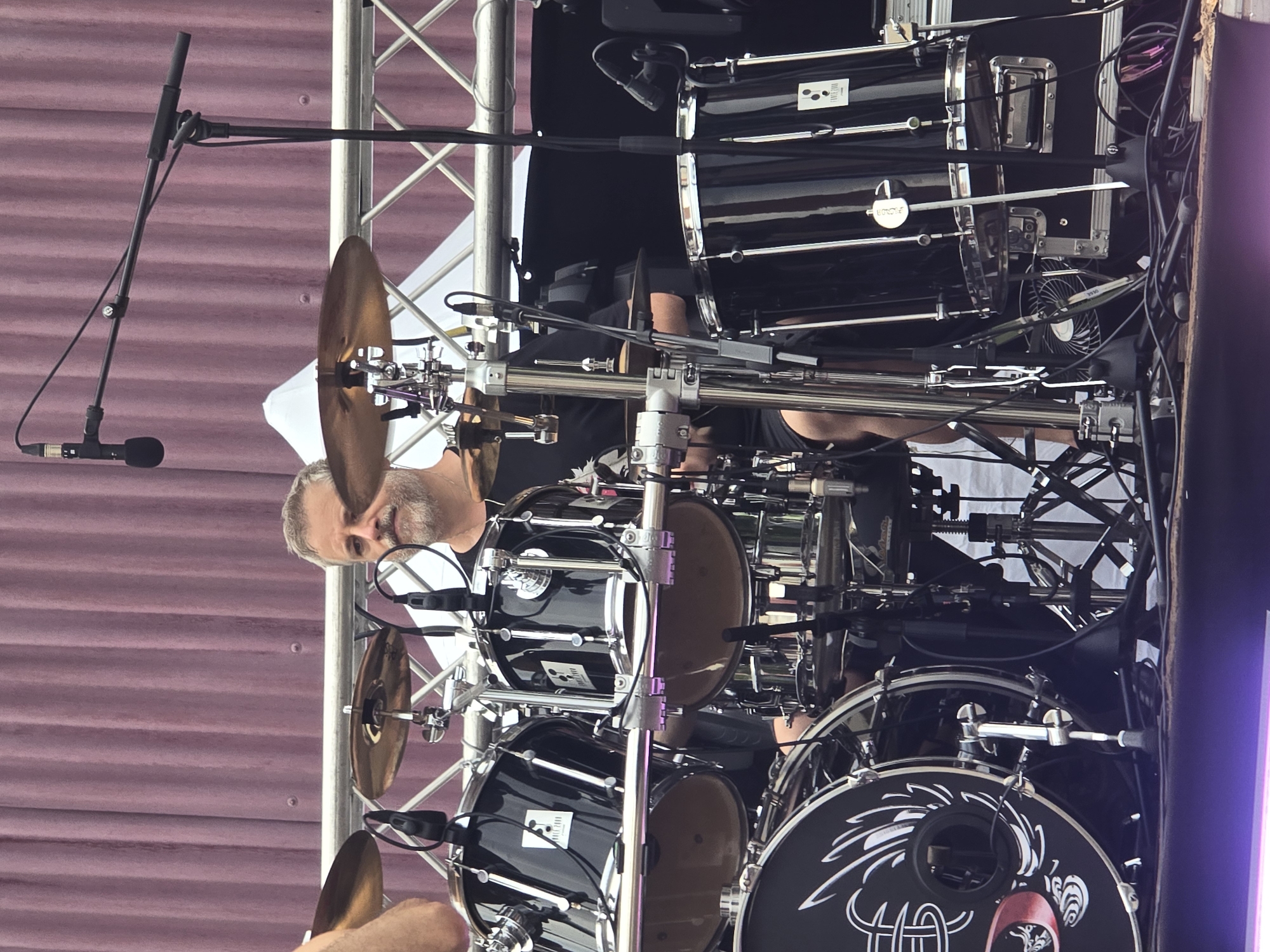 The image shows a male drummer with graying hair and a beard playing a drum set on a stage. The drummer is wearing a black t-shirt and is positioned behind a complex arrangement of drums and cymbals. Various microphones are positioned to capture the sound of the drums. In the background, a purple curtain-like material hangs, and a metal truss structure supports some of the equipment. The stage appears to be outdoors, with a hint of a brighter, possibly overcast sky or stage lighting at the bottom of the frame. The drummer is actively engaged in playing. The specific location in Cornellà de Llobregat, Spain, is not discernible from the image itself.