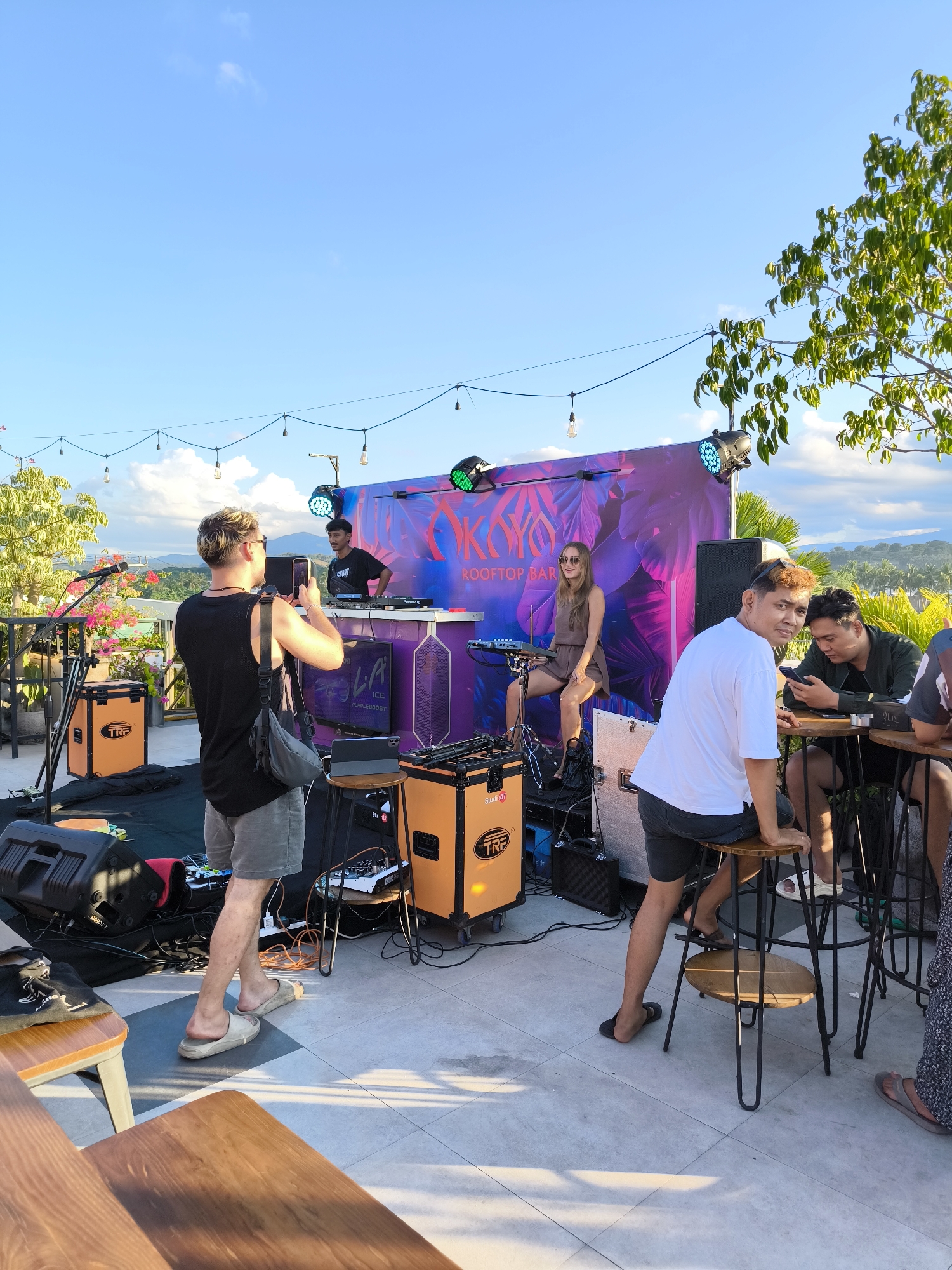 The image depicts an outdoor event, likely a rooftop bar in Sumbawa Besar, Indonesia, as suggested by the backdrop bearing the name "OKYO Rooftop Bar." A DJ is positioned at a platform behind a woman seated on a stool playing a keyboard. A man in the foreground is taking pictures of the band. Several other people are seated on stools at a table, some looking at their phones. There are speakers and other musical equipment on the stage. The sky is blue with some clouds, and string lights are hung overhead. Some foliage is also visible on the right side of the frame.