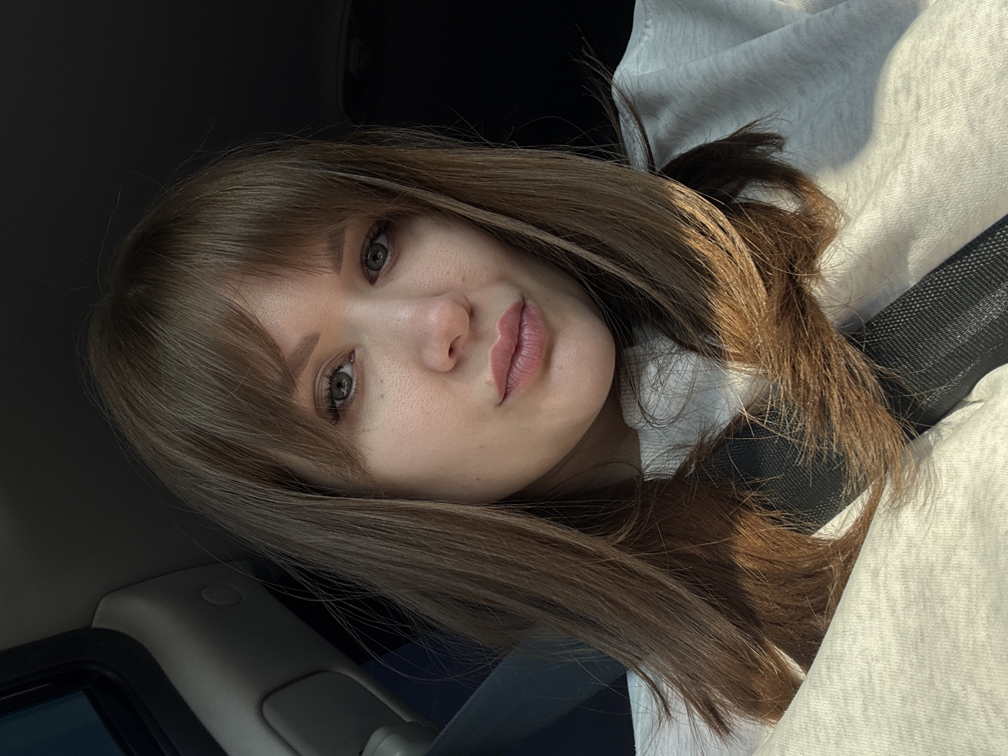 A close-up, selfie-style photograph depicts a young woman with brown hair and bangs. She is wearing a light-colored sweatshirt and a seatbelt. The background suggests she is inside a vehicle, possibly a car. The environment is not detailed enough to determine her location as Alnashi, Russia.
