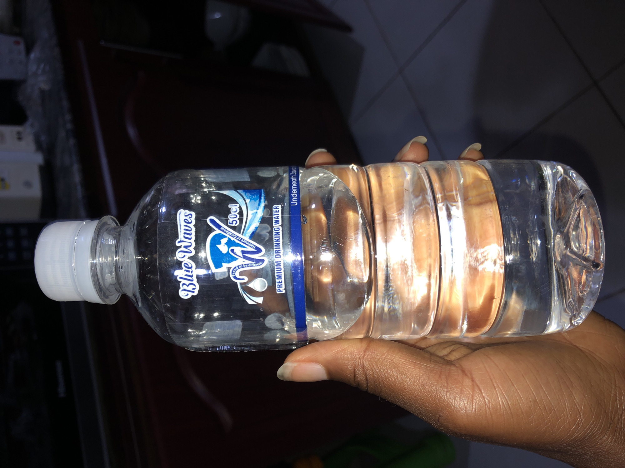 A person is holding a clear plastic bottle of Blue Waves premium drinking water. The bottle is labeled with the brand name "Blue Waves" in a blue script font and a blue wave graphic. Text on the label also reads "50cl" and "PREMIUM DRINKING WATER". Underneath "Blue Waves" there is smaller text that says "Hallmark of Excellence. Need to be different". The water inside the bottle is clear. The person's hand, with manicured fingernails, is supporting the bottle. The background is dark and out of focus, but suggests an indoor setting with some reflective surfaces. The lighting is dim. The overall impression is of a casual presentation of a product.
