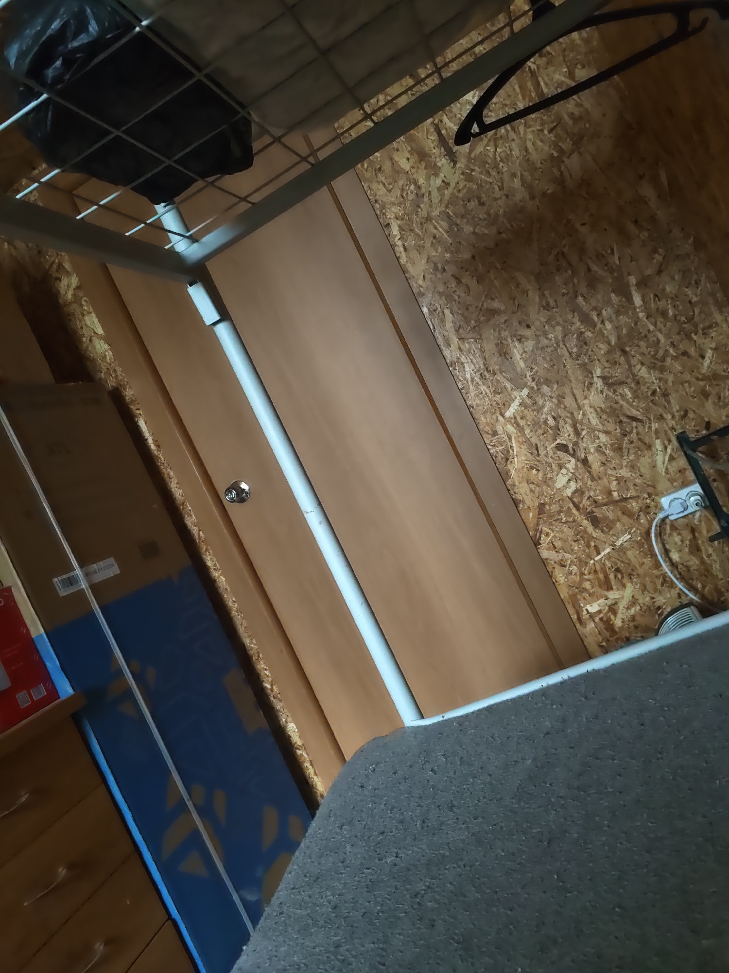 The image depicts an indoor scene, likely a storage area or unfinished room, with walls made of Oriented Strand Board (OSB). A metal bed frame is visible at the top of the image, with a black plastic bag placed on its mesh base. A black plastic clothes hanger hangs from a metal rail on the right side. To the left, a tall, light-brown wooden door with a silver doorknob is partially visible. In front of the door, there are several cardboard boxes, one of which is open and contains blue and brown packaging. A drawer unit with three visible drawers is situated in the foreground on the left, and a section of dark gray, textured material, possibly a mattress or carpet, fills the lower right portion of the frame. A white electrical cord is plugged into a wall socket on the OSB wall. The scene is dimly lit, suggesting it might be a closet, attic, or basement in a building located in Платошино, Russia. There are no people or animals visible in the image.