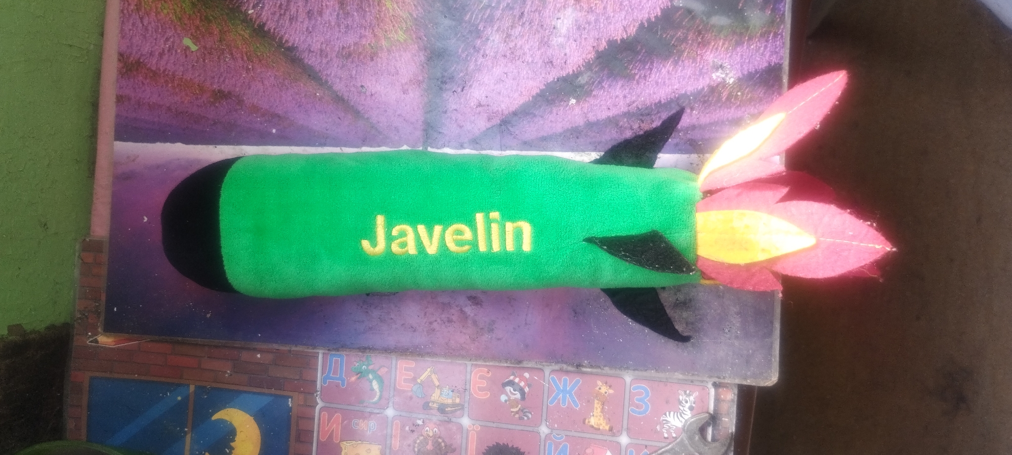 A plush toy rocket is the central subject of this image, lying horizontally. The rocket is primarily green, with a black tip. The word "Javelin" is embroidered in yellow thread across its body. At the rear of the rocket, colorful fabric shapes resembling flames or exhaust are attached, predominantly in shades of pink and yellow. Black fabric fins are also visible on the rocket. The rocket appears to be resting on a surface with a patterned background. The pattern includes what looks like a brick wall motif along the bottom edge and a larger, abstract design above it, with shades of purple and pink reminiscent of a lavender field. Below the rocket, on the patterned surface, there are what appear to be alphabet tiles with cartoon images, including a dragon, a construction vehicle, a raccoon, a giraffe, and a zebra. The image is taken from a top-down perspective, with some parts of the background out of focus. The overall lighting suggests it might be daytime. The setting appears to be outdoors or in an area with natural light, possibly a yard or balcony, given the rough textures and the presence of what looks like a brick wall. No people are visible in the frame. The location context of Zaporizhzhia, Ukraine, is provided but not visually evident from the image itself.